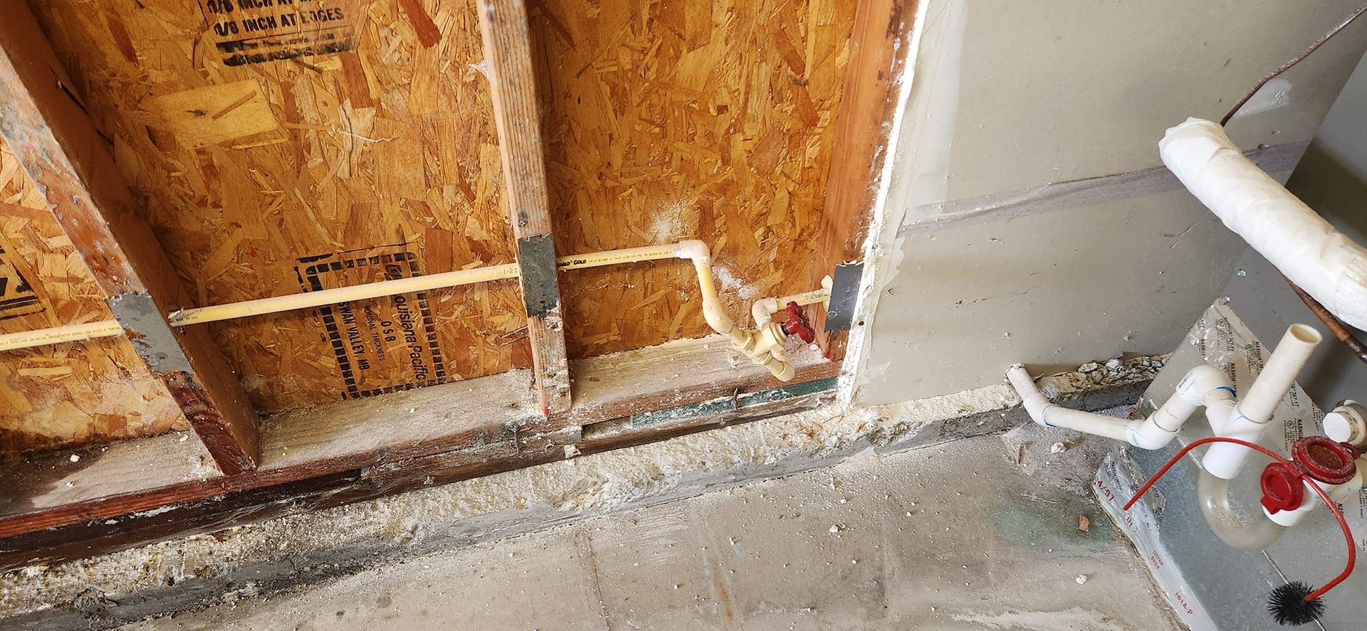 An unfinished interior wall showing exposed wooden studs, spray foam, and plumbing lines near a concrete floor.