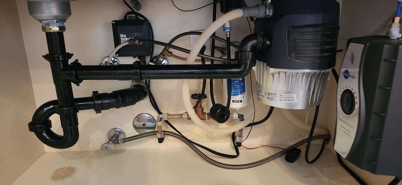Under-sink plumbing showing a black p-trap connected to a garbage disposal and an electric water heater unit.