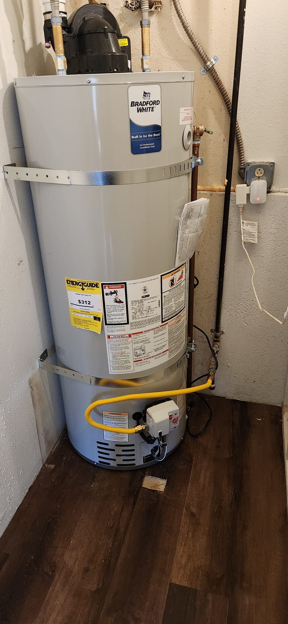 A gray residential water heater stands in a corner on a dark wood-look floor, secured to the wall with metal straps.