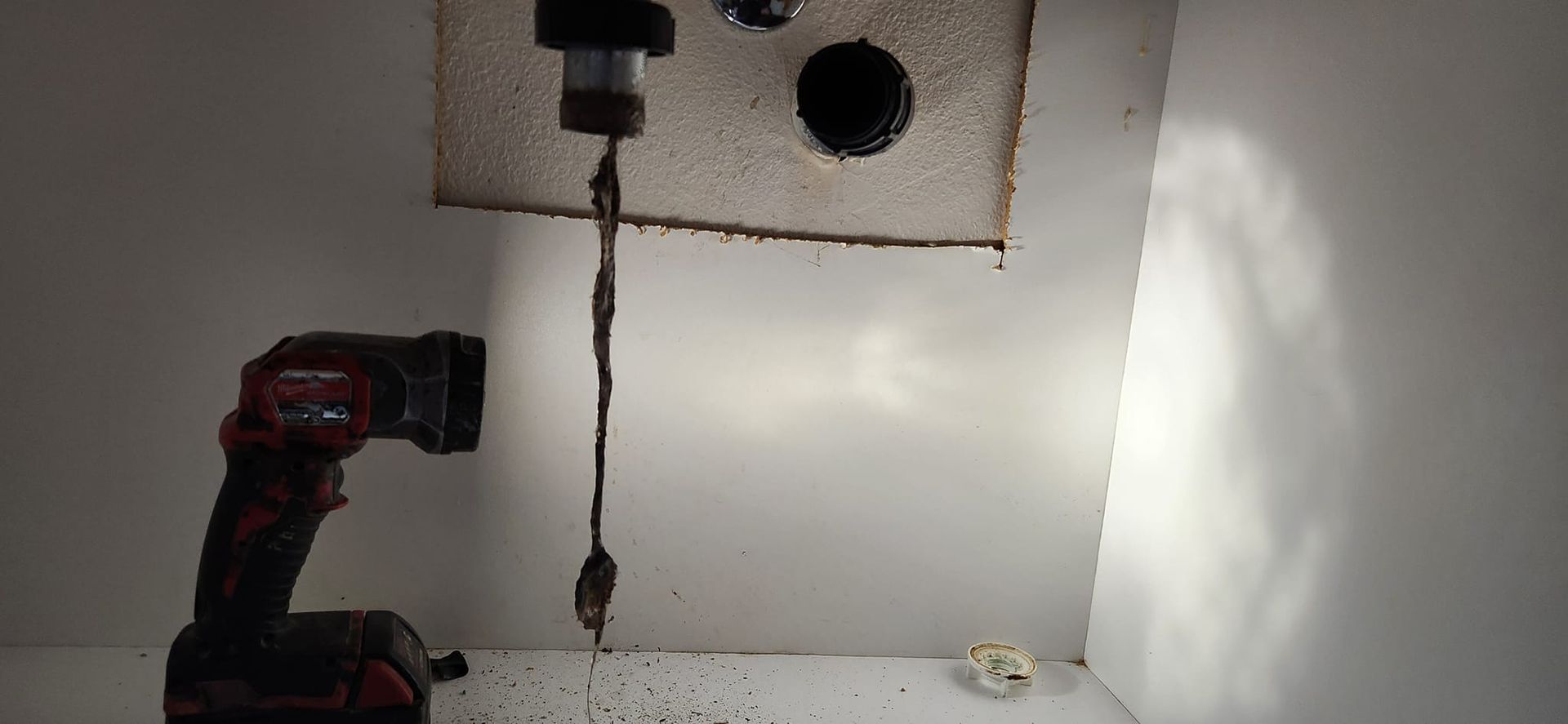 A handheld work light illuminates a long, dark clog being pulled from an open plumbing pipe in a wall.