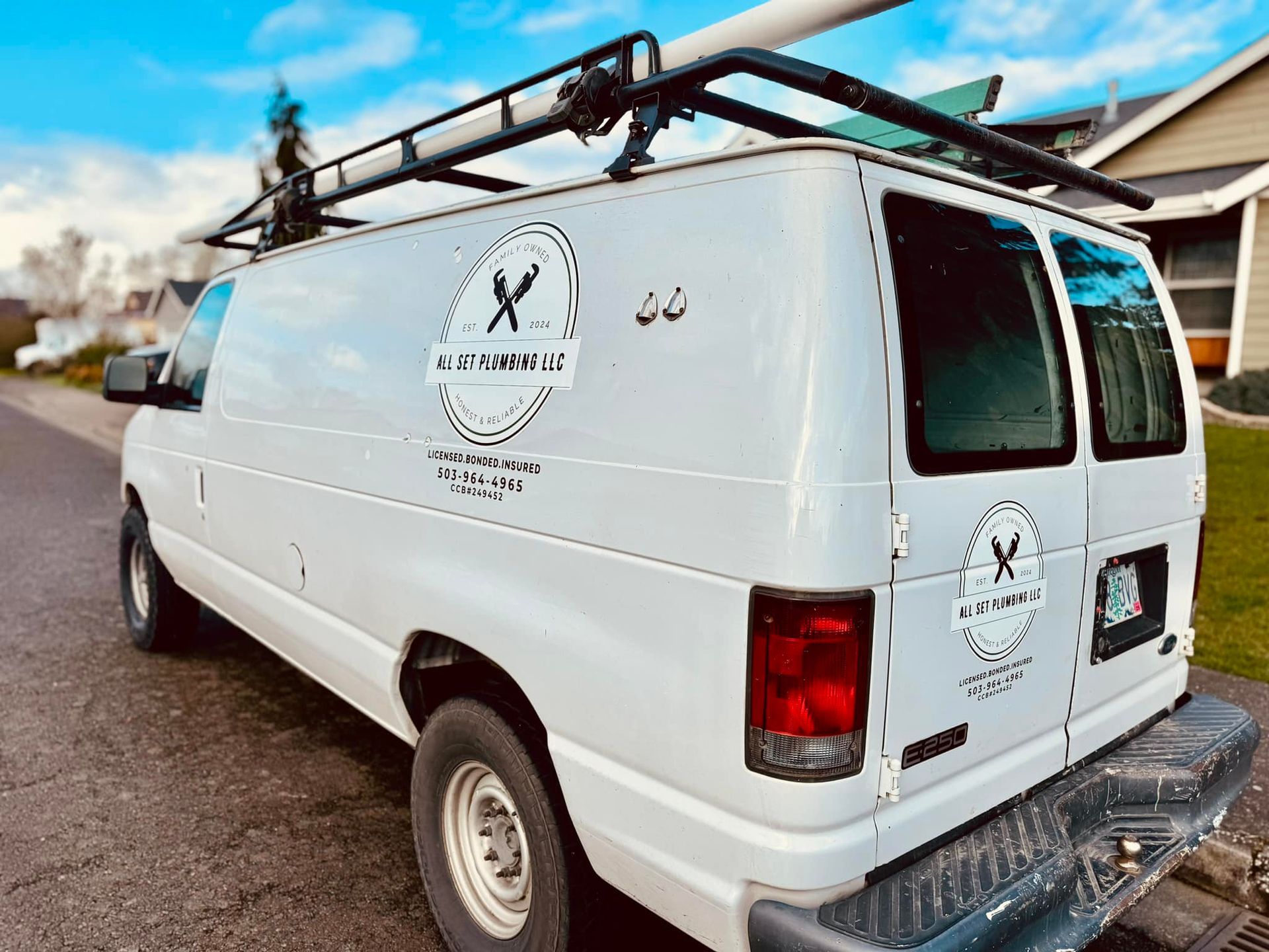 A white service van parked on a residential street with plumbing company logos on the side and rear doors.