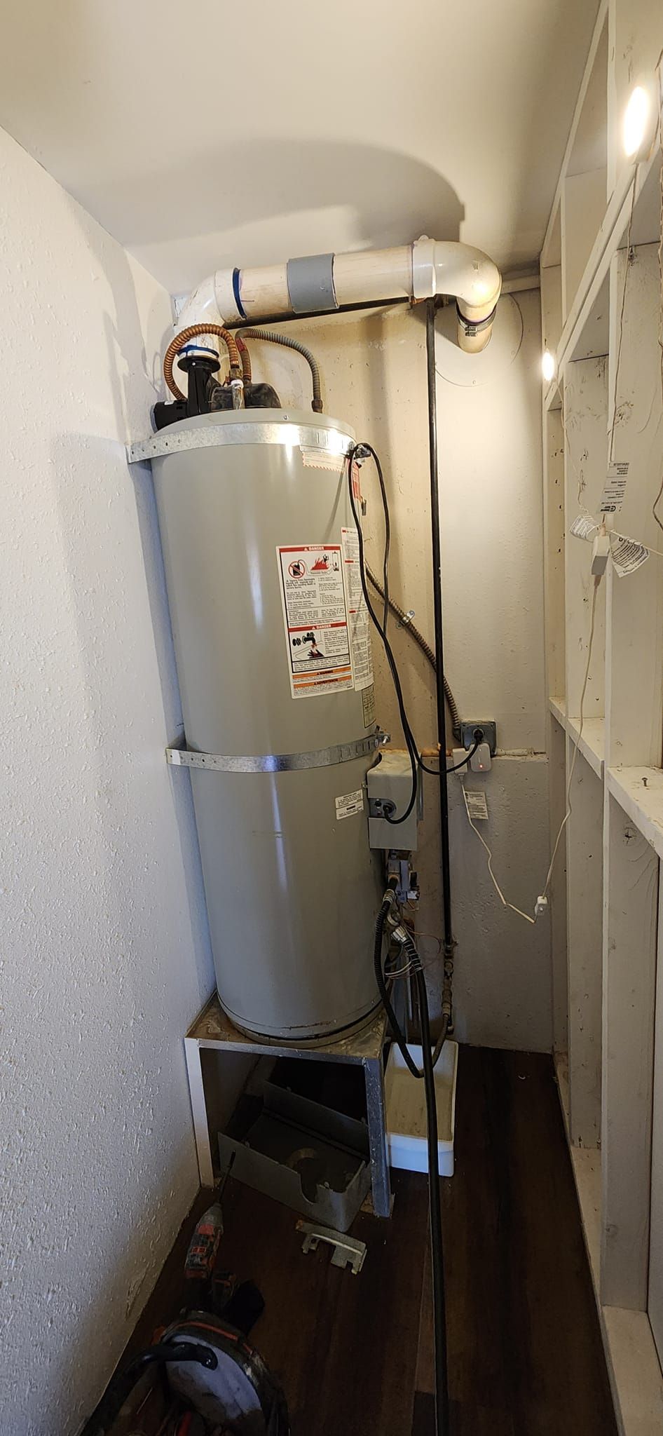 A tall, gray water heater sits on a wooden platform inside a closet, with a white exhaust pipe extending from the top.