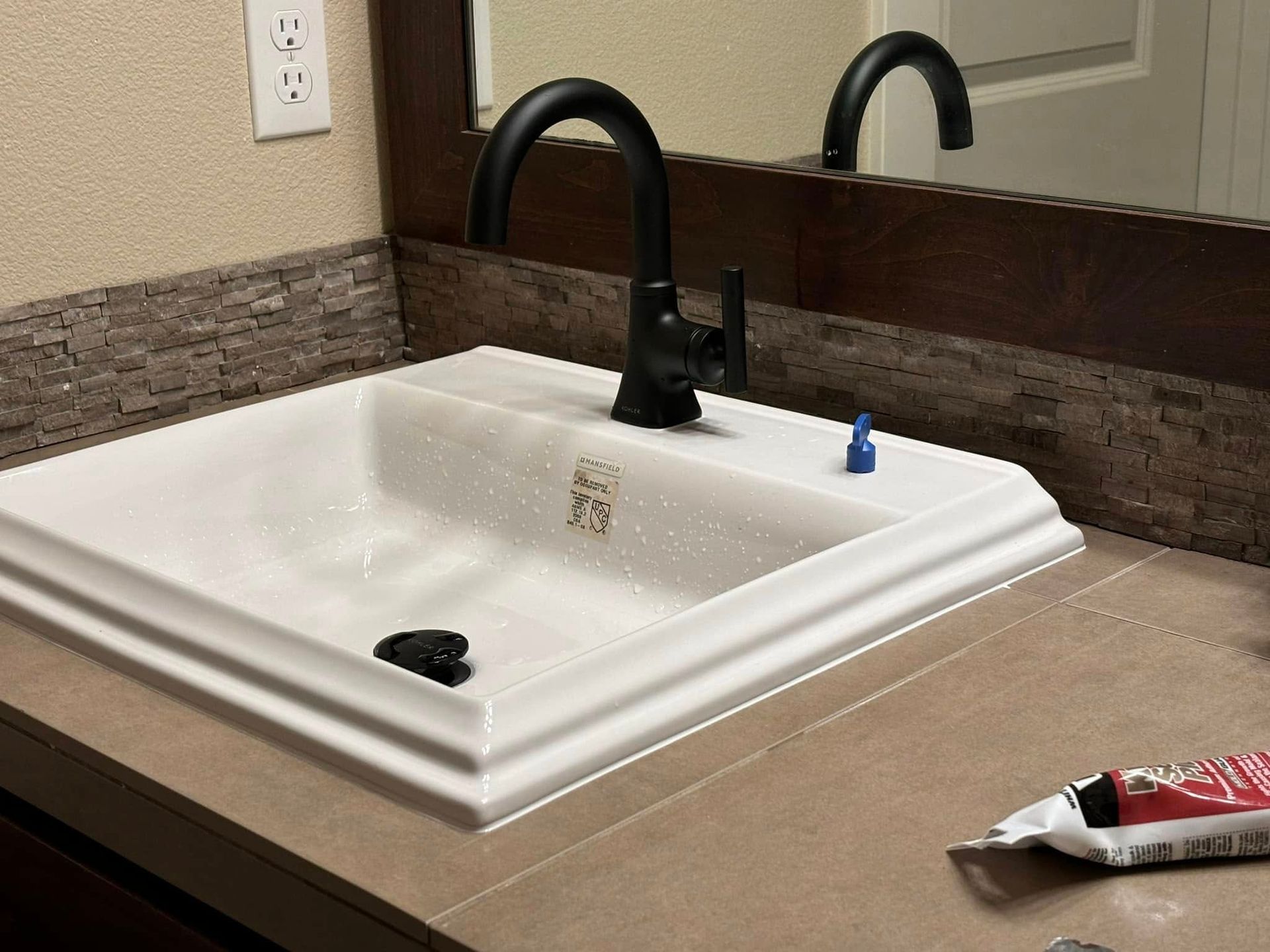 A modern white bathroom sink with a black faucet on a tiled countertop, next to a tube of toothpaste.