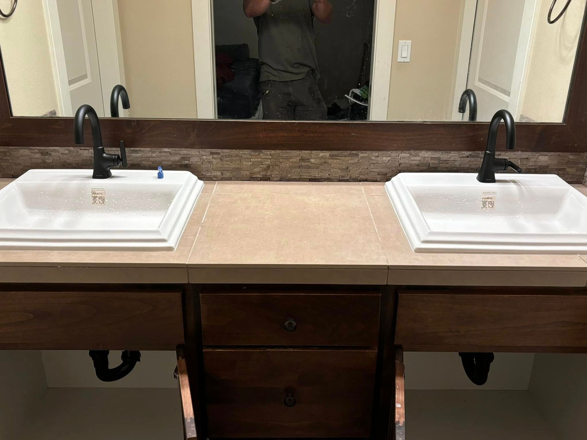 A double bathroom vanity with white rectangular sinks, black faucets, and a wood-framed mirror in a tan room.