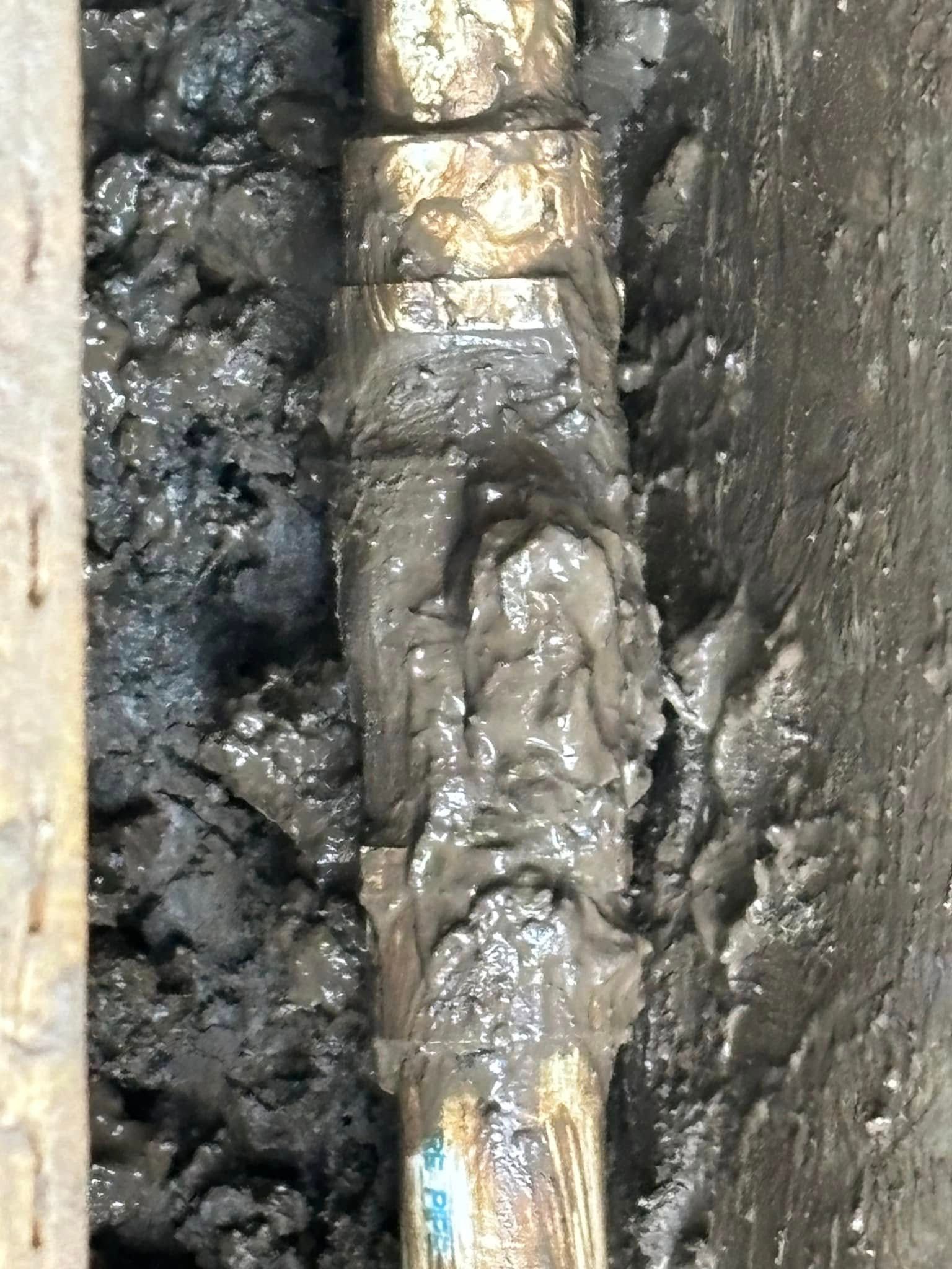 A muddy, metallic plumbing connection or pipe coupling is partially buried in dark, wet soil.