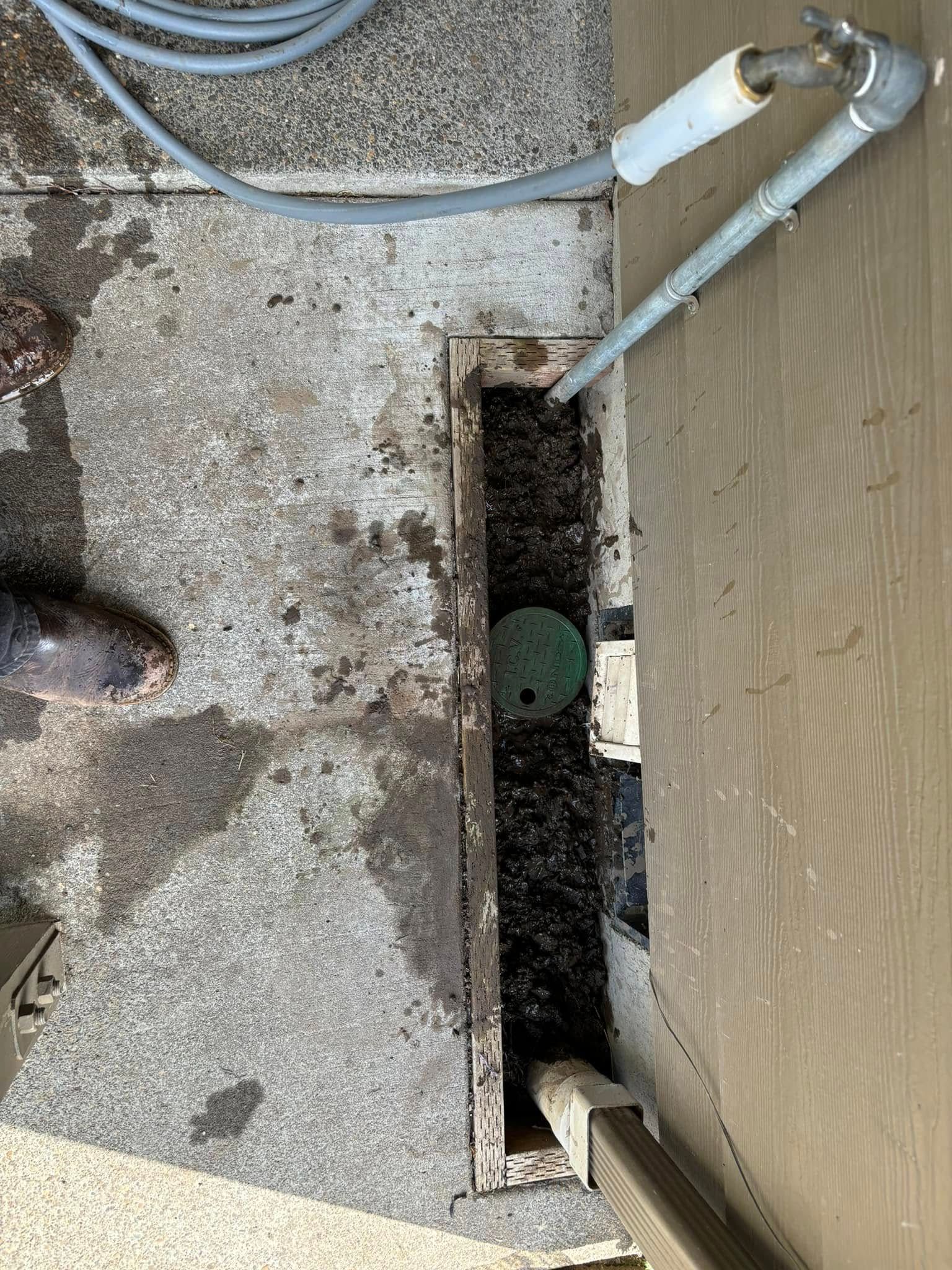 An outdoor narrow trench near a building wall containing a round green valve box cover next to a hose spigot.