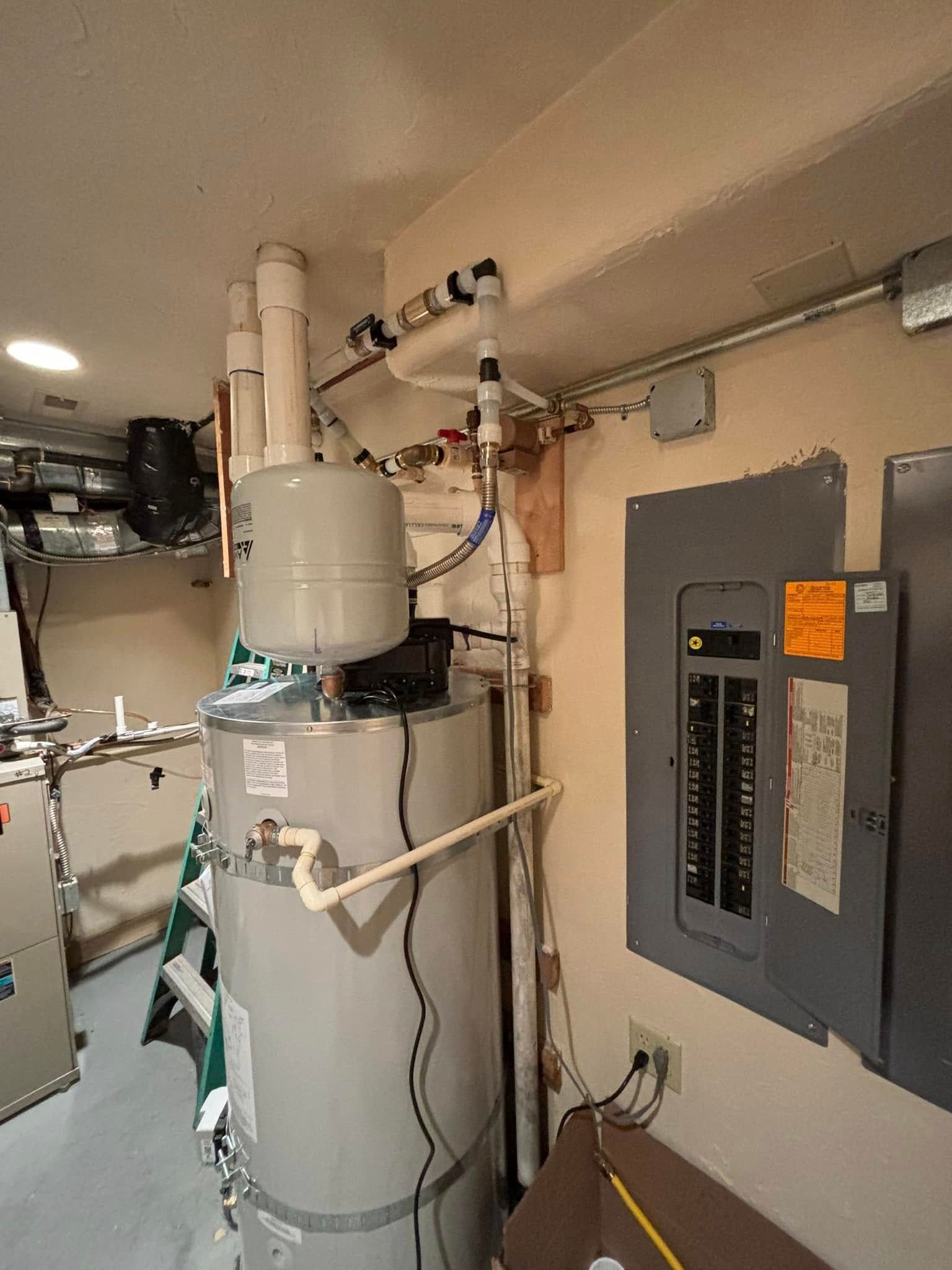 An electric water heater with an expansion tank next to an electrical panel in a room with white walls.