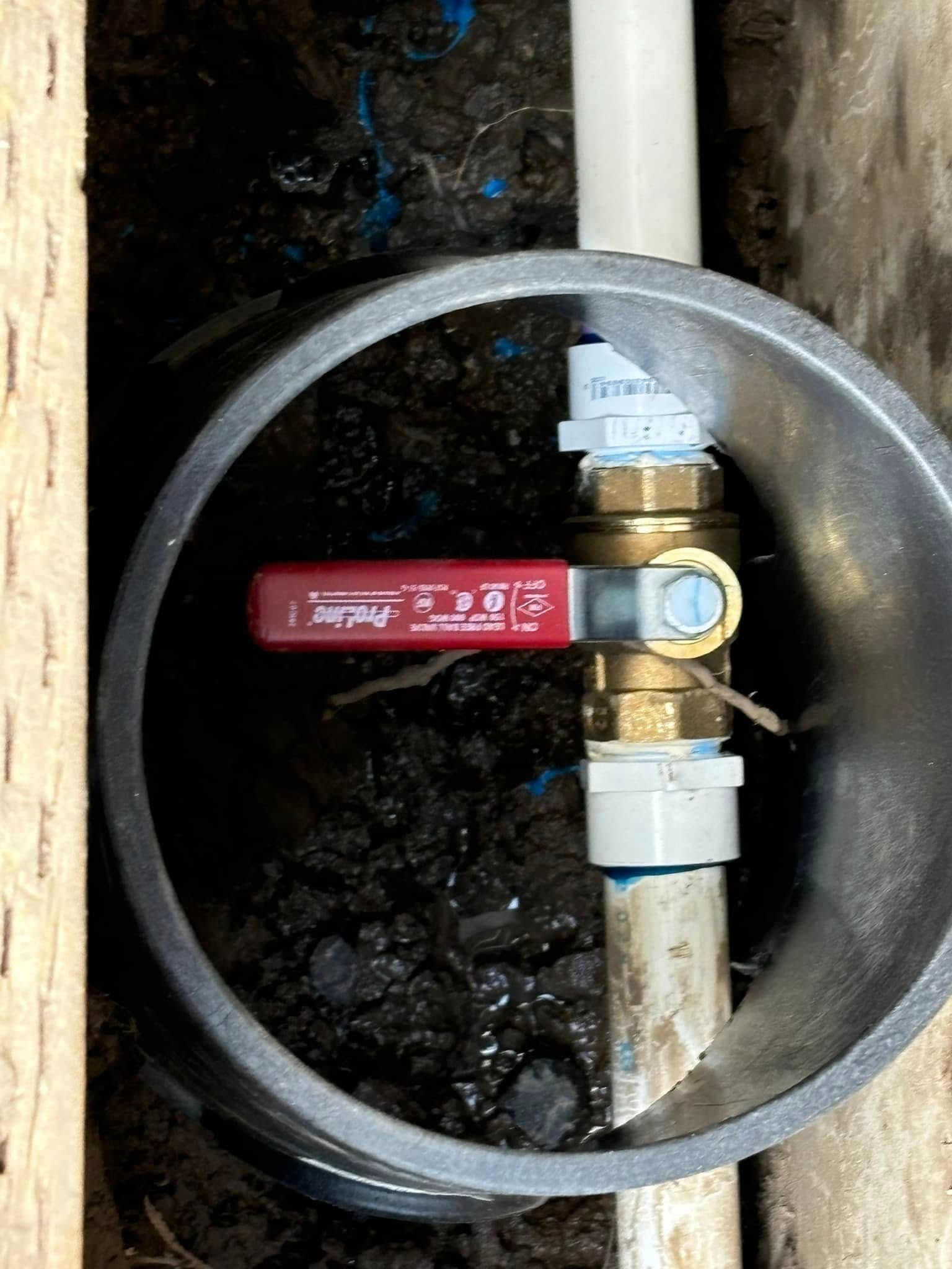 A brass ball valve with a red handle installed on a white PVC pipe inside a circular grey housing sleeve in the ground.