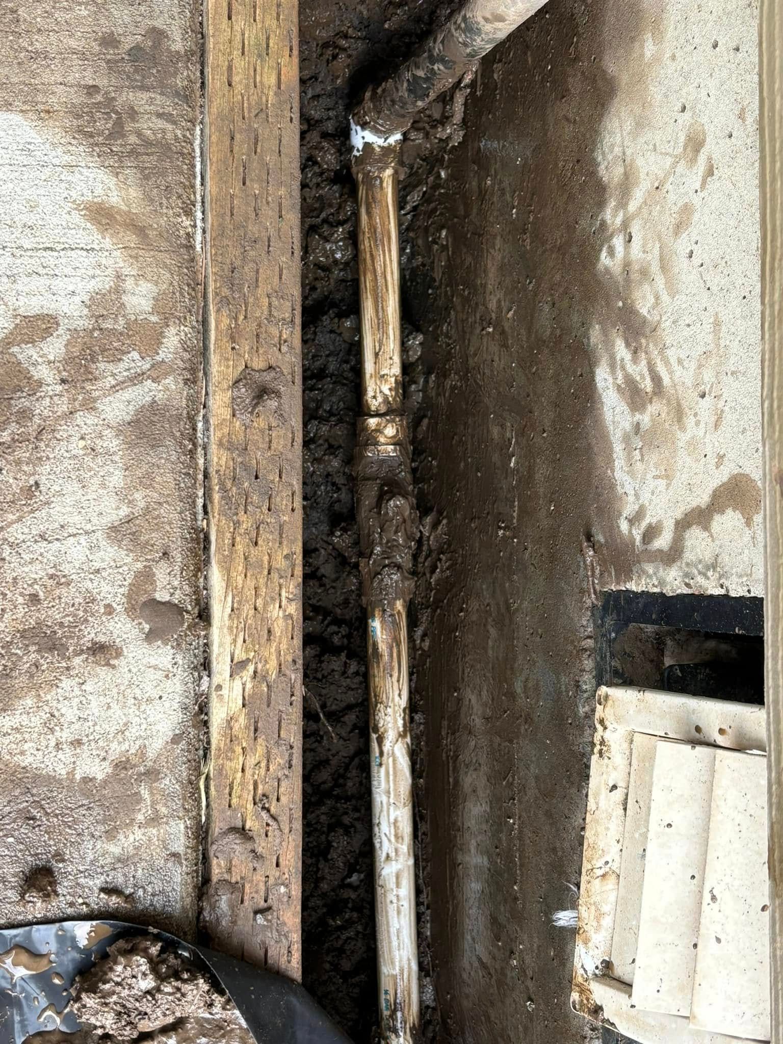 A light-colored pipe lies in a narrow dirt trench between a wooden board and a concrete foundation.