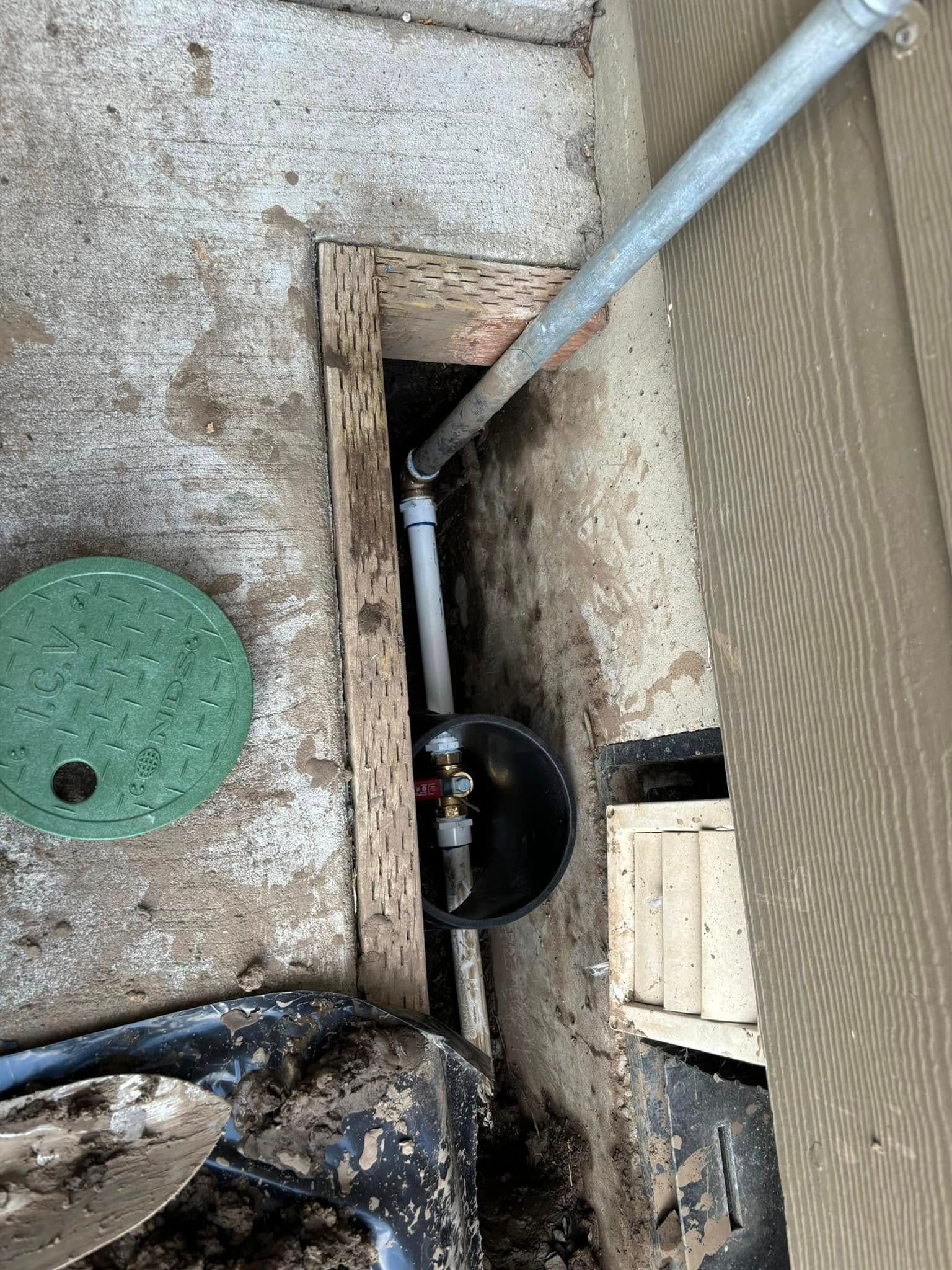A pipe connection inside a rectangular trench cut into concrete next to a building wall, with a green circular cover nearby.