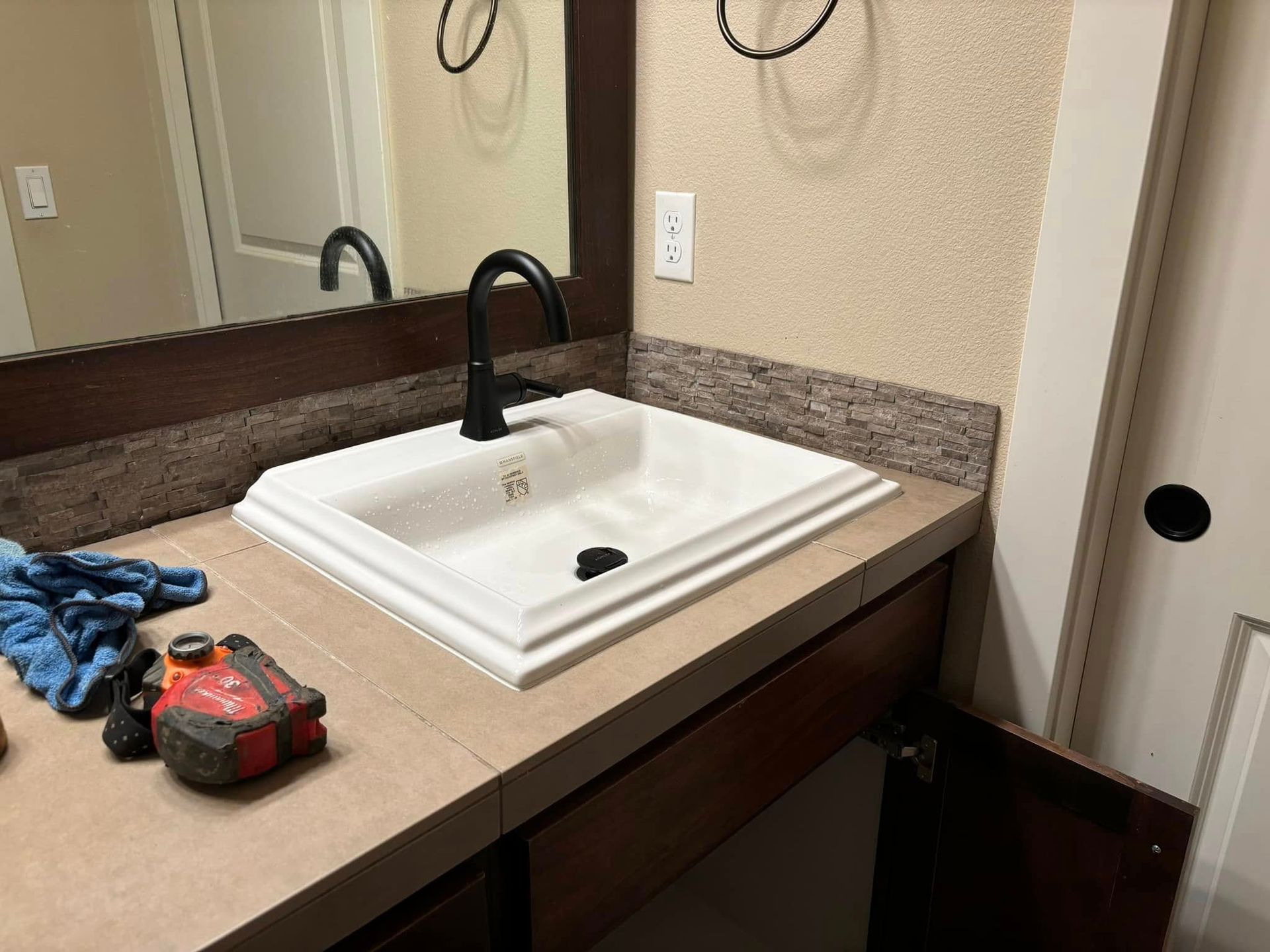 A modern bathroom sink with a white rectangular basin and a black faucet, set in a tiled vanity with tools on the side.