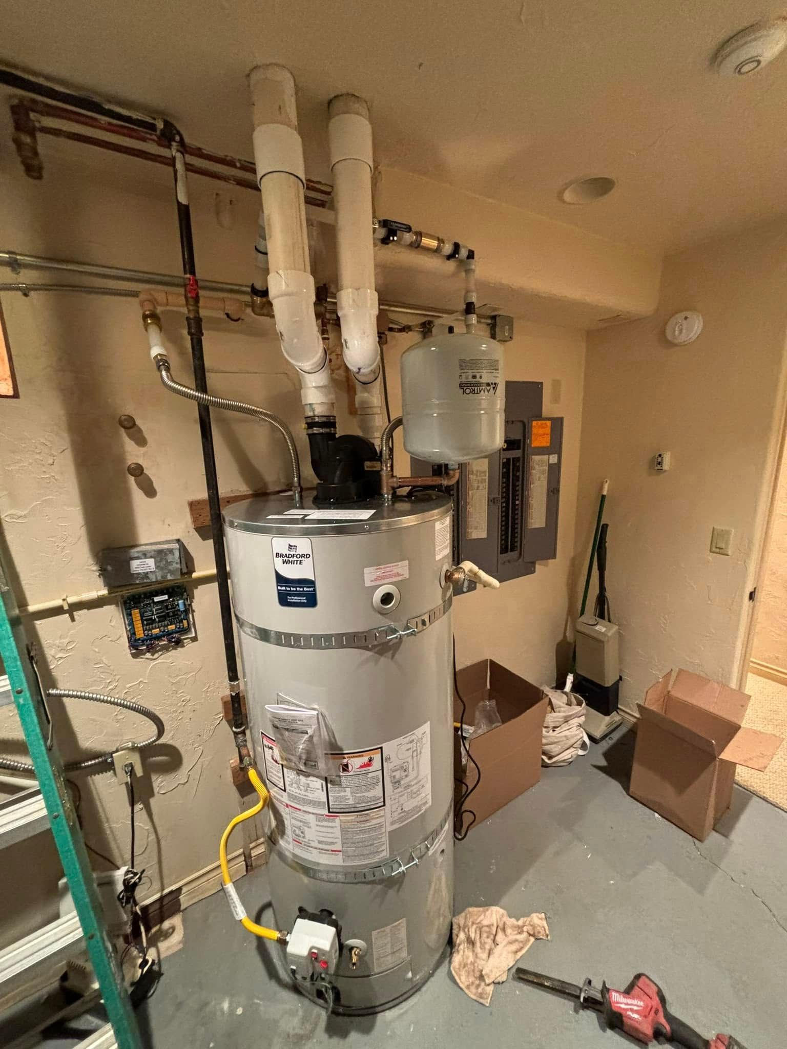 A grey residential water heater with two white PVC exhaust pipes, a pressure tank, and tools in a basement utility room.