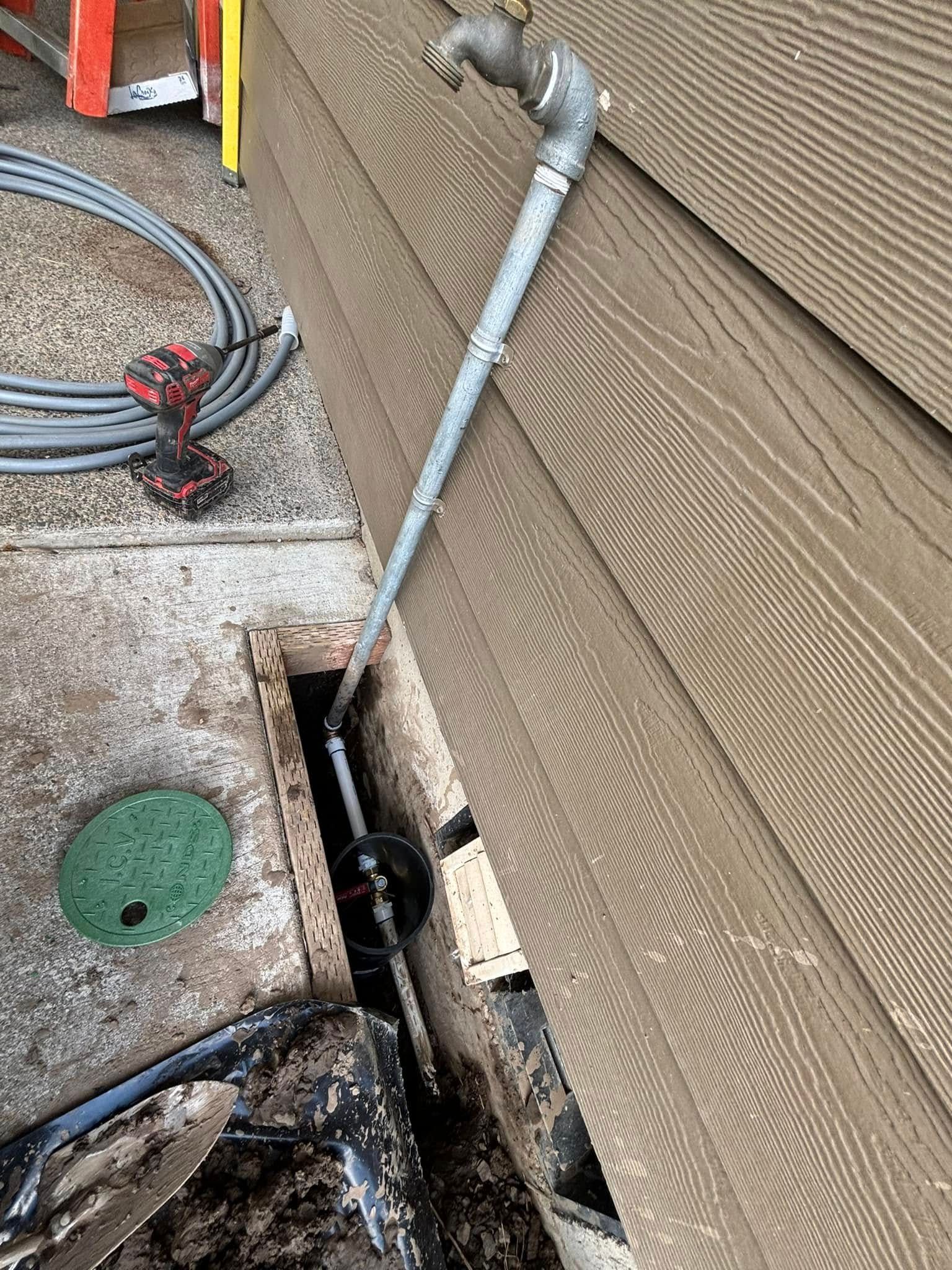 A galvanized metal pipe riser with an outdoor faucet emerges from an open trench against the side of a house.