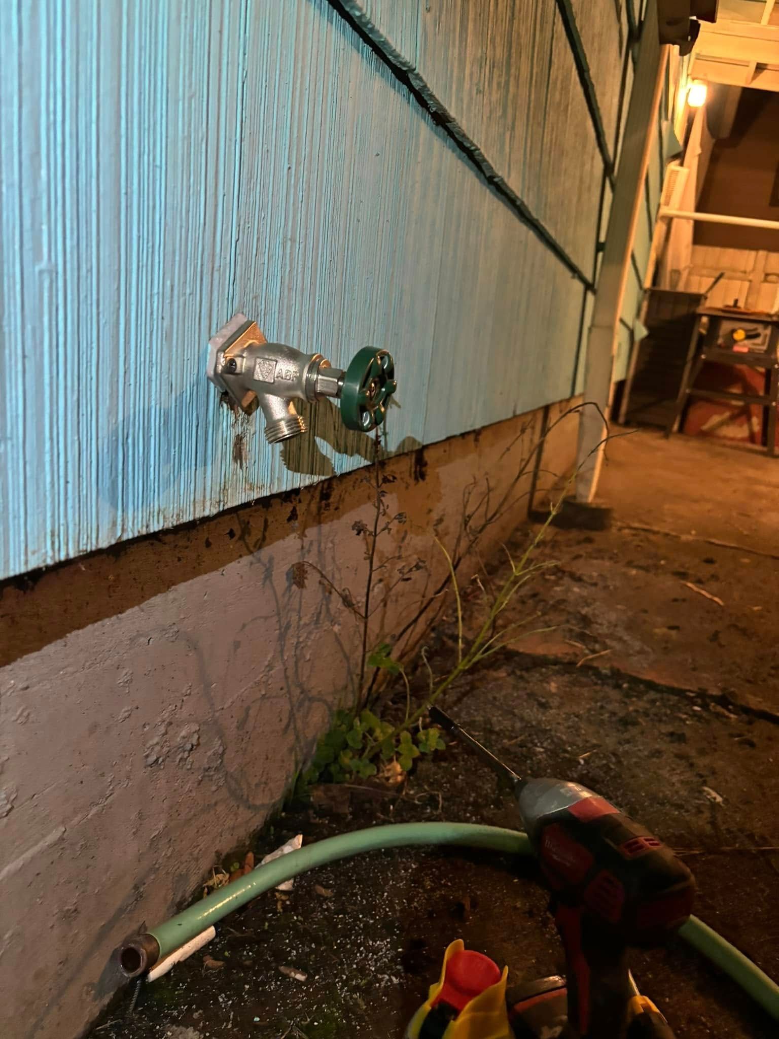 A green-handled outdoor water spigot mounted on blue siding above a concrete foundation with a hose lying on the ground.