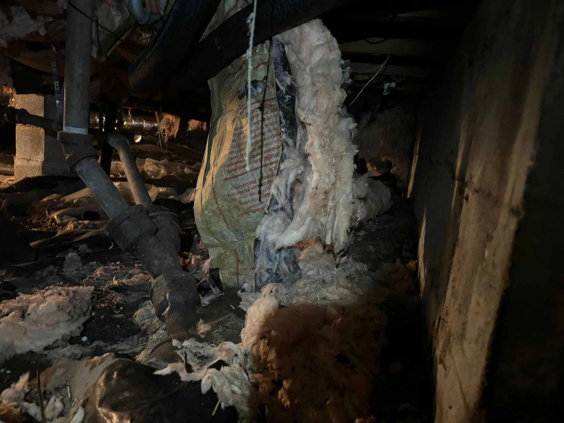 A dark, damp crawl space with hanging, damaged insulation and exposed pipes near a concrete foundation wall.