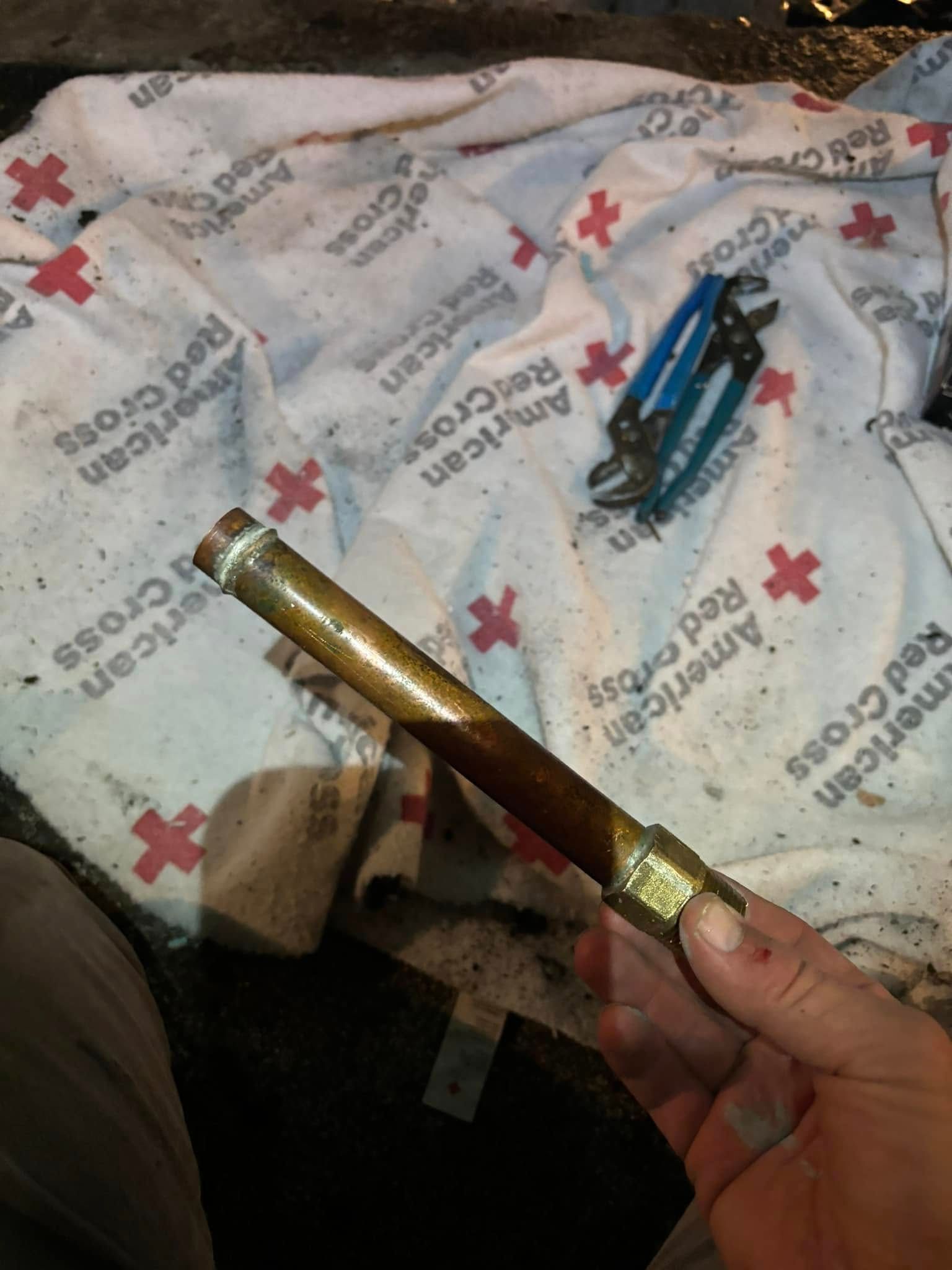 A hand holds a brass pipe fitting over a white American Red Cross blanket, with blue pliers in the background.