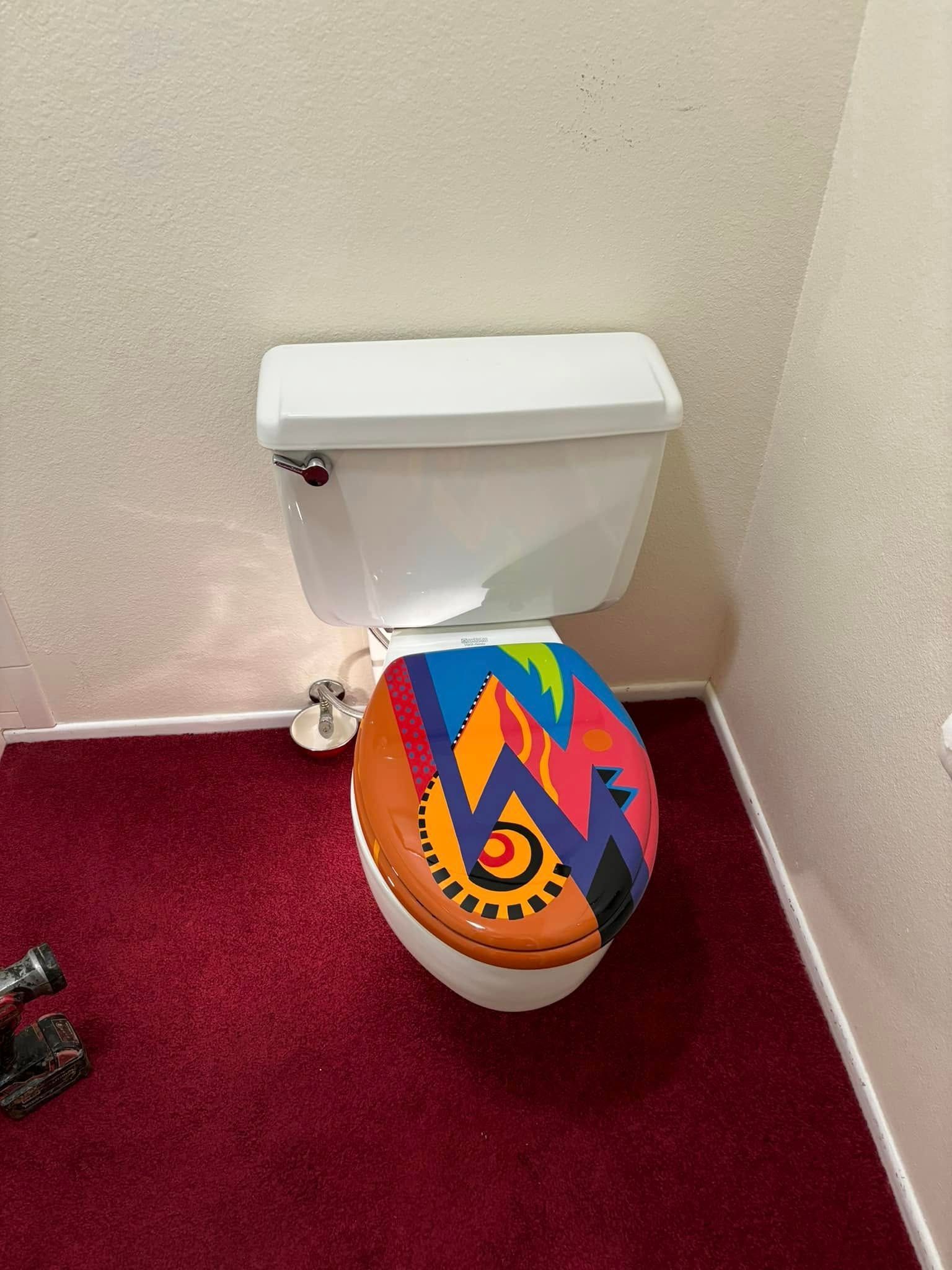 A white toilet with a brightly colored, abstract patterned seat, positioned on a deep red carpeted floor.