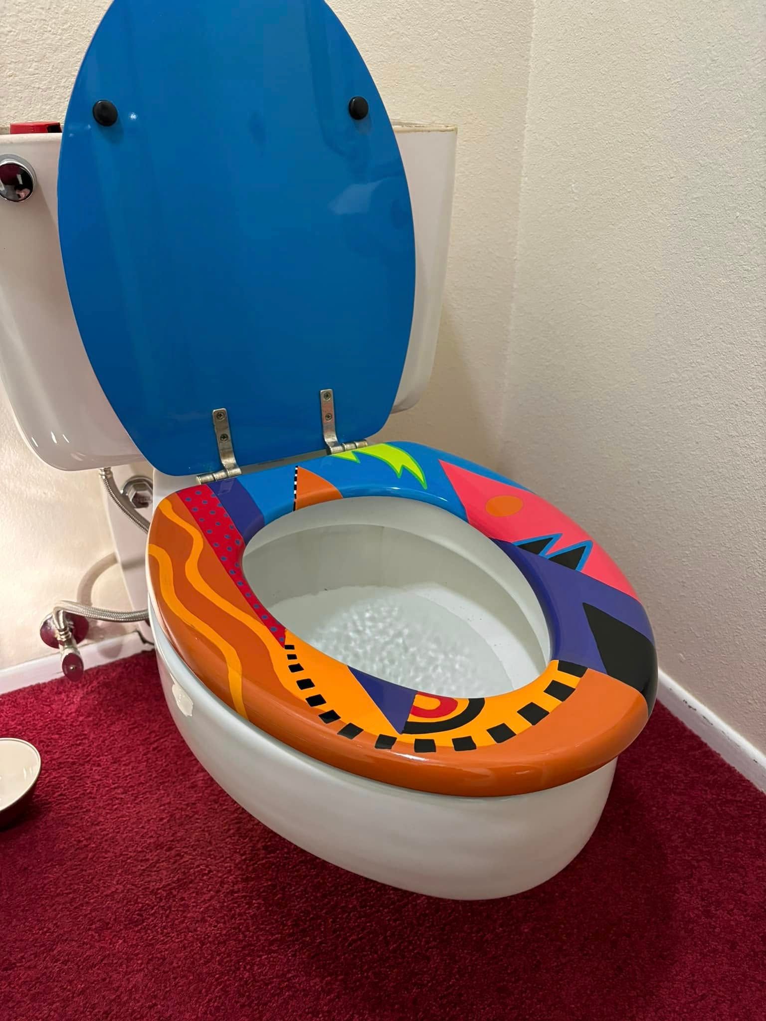 A white toilet with a bright blue lid and a multicolored, patterned seat sitting on a red carpet.