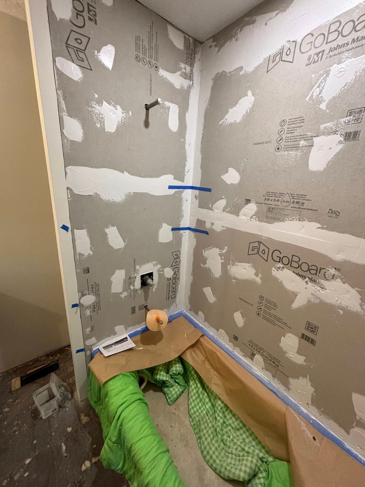 A shower alcove with GoBoard panels installed, featuring blue tape at the seams, a faucet valve, and a protective tub cover.