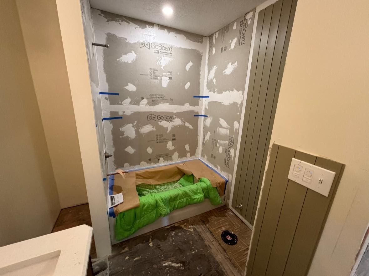 A bathroom under construction showing a bathtub alcove with unfinished drywall, blue tape, and protective green sheeting.