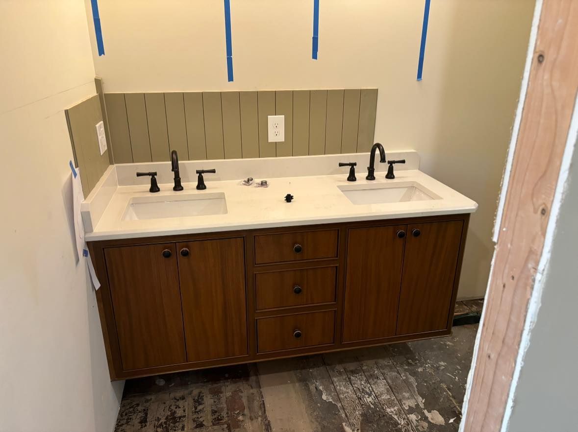 A double-sink bathroom vanity with wood cabinets and a white countertop, under construction in a room with unfinished walls.
