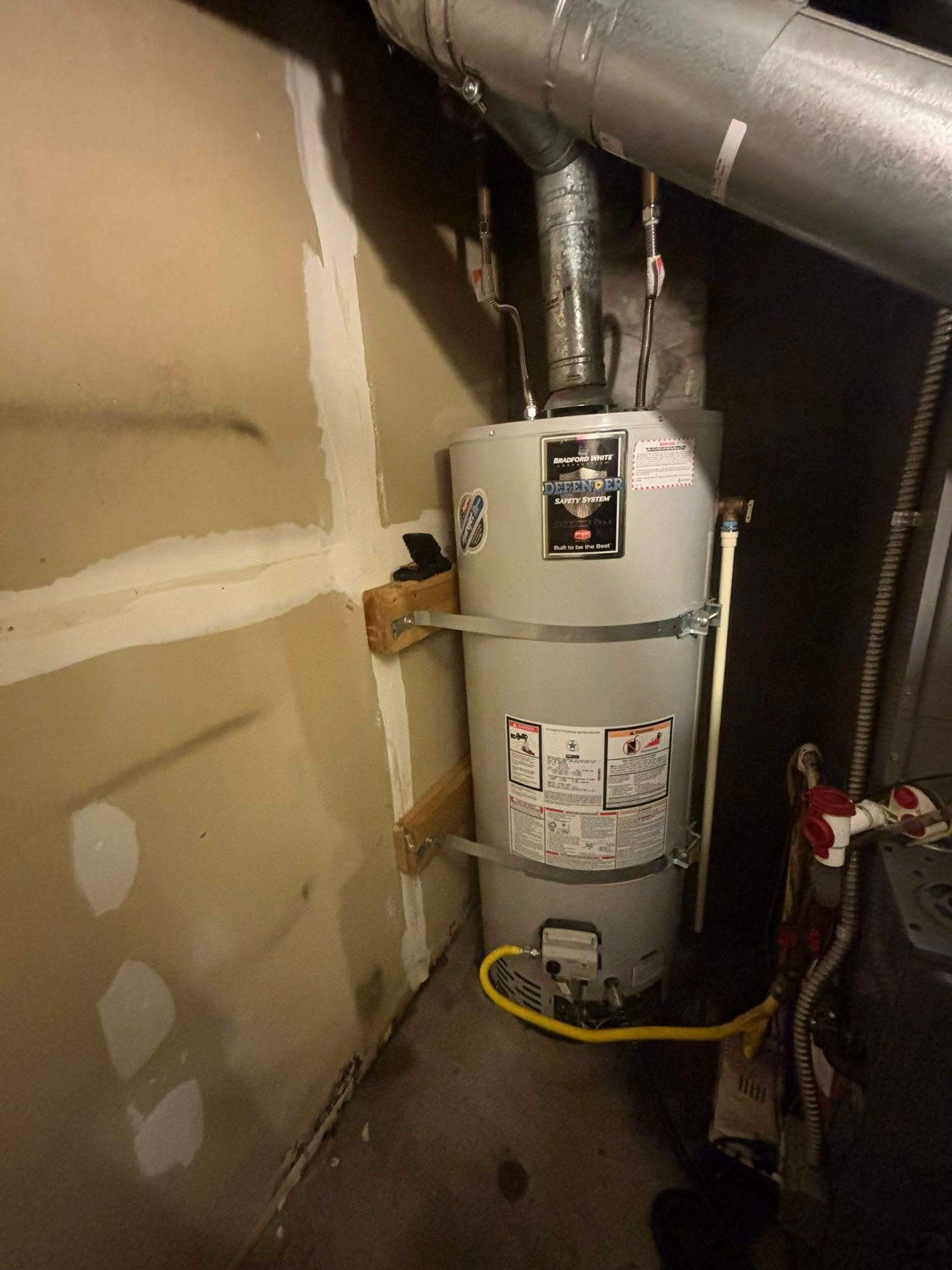 A residential gas water heater, secured to a drywall wall with metal straps and wooden blocks, connected to vent piping.