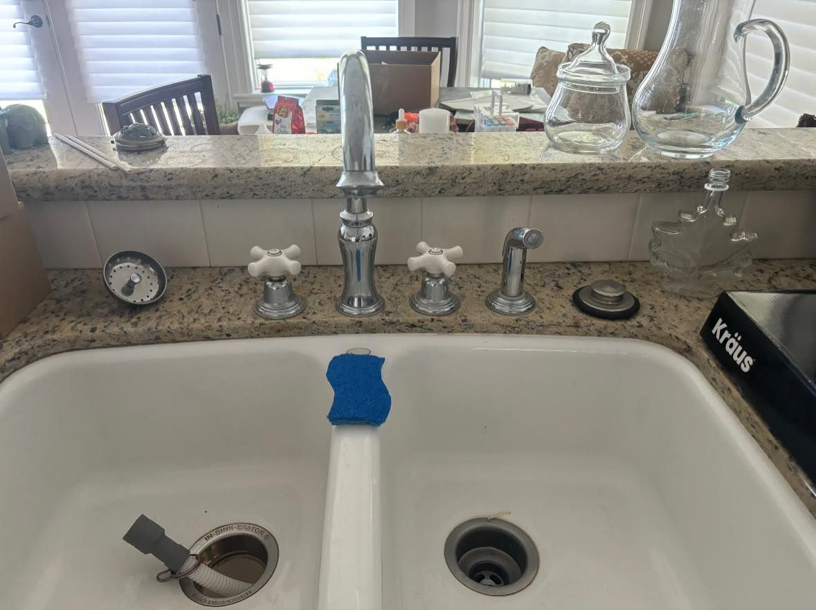 A white double-basin kitchen sink with a chrome faucet, knobs, and a blue sponge resting on the center divider.