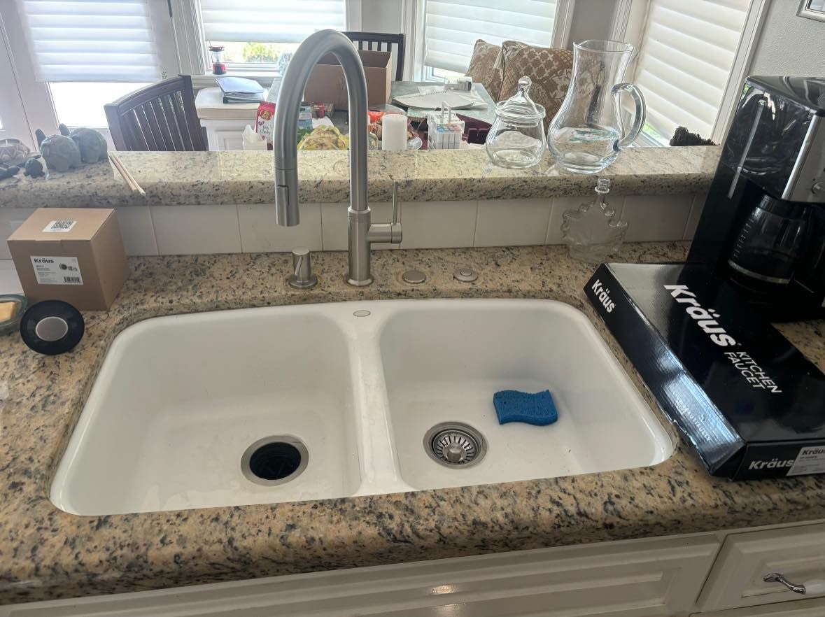 A double-basin white kitchen sink with a metal faucet on a granite countertop, with kitchen items surrounding it.