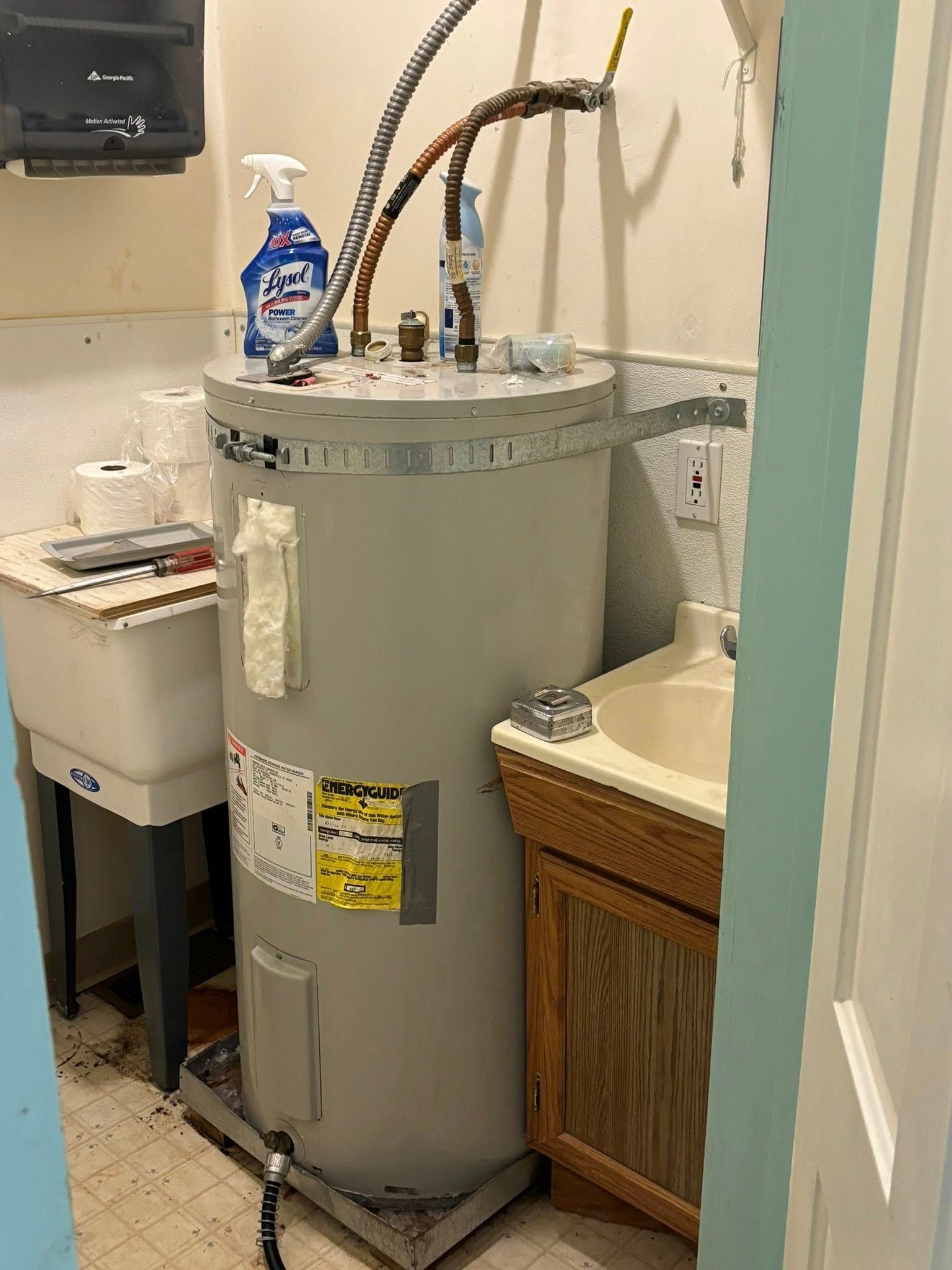 A utility room with a grey water heater positioned between a white laundry sink and a small wooden bathroom vanity.
