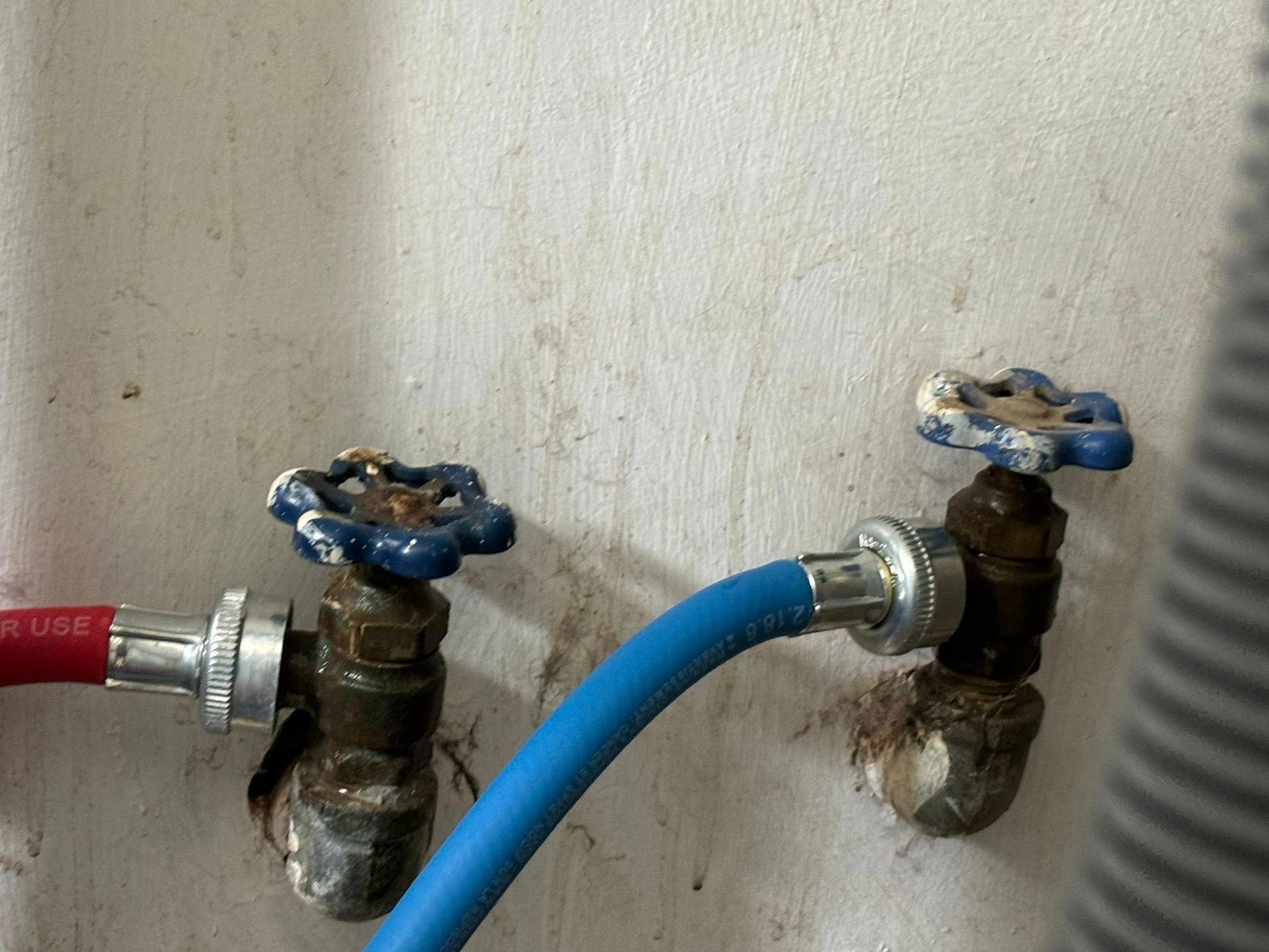 Two metal water valves with blue handles attached to red and blue hoses against a white wall.
