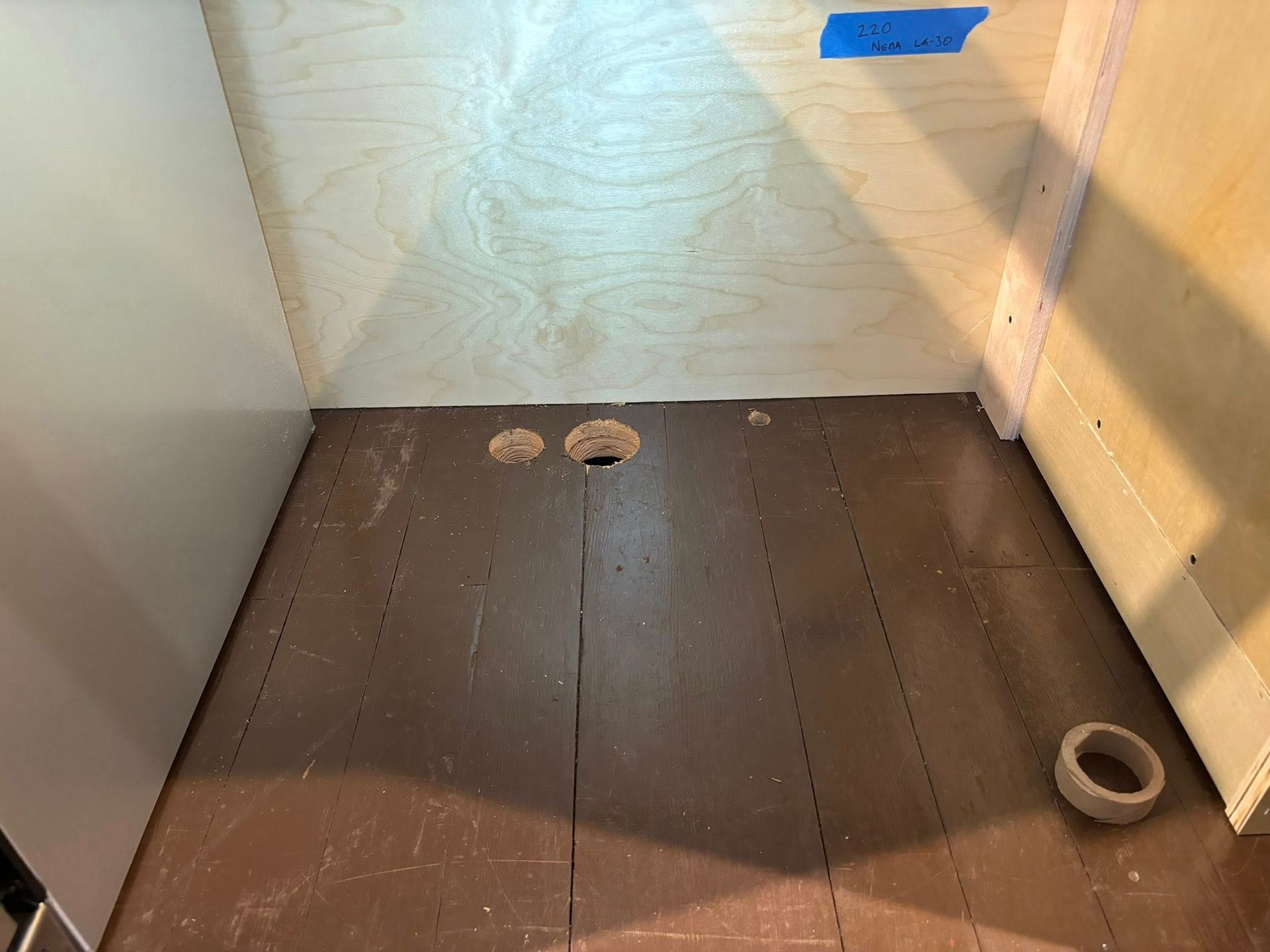 The interior of a wooden cabinet base with two holes for plumbing and a strip of blue painter's tape on the back wall.
