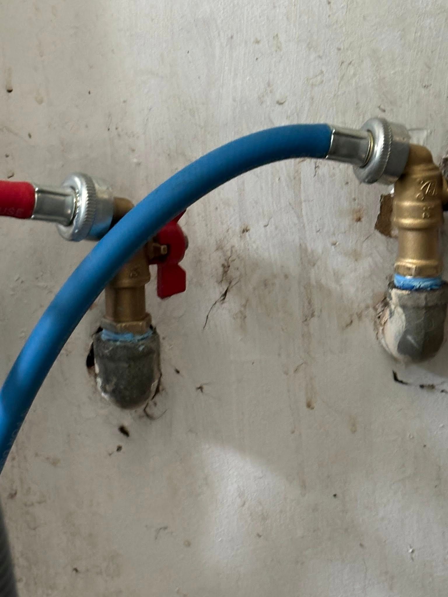 A blue flexible hose connected to a brass plumbing valve and fitting on a textured, off-white wall.