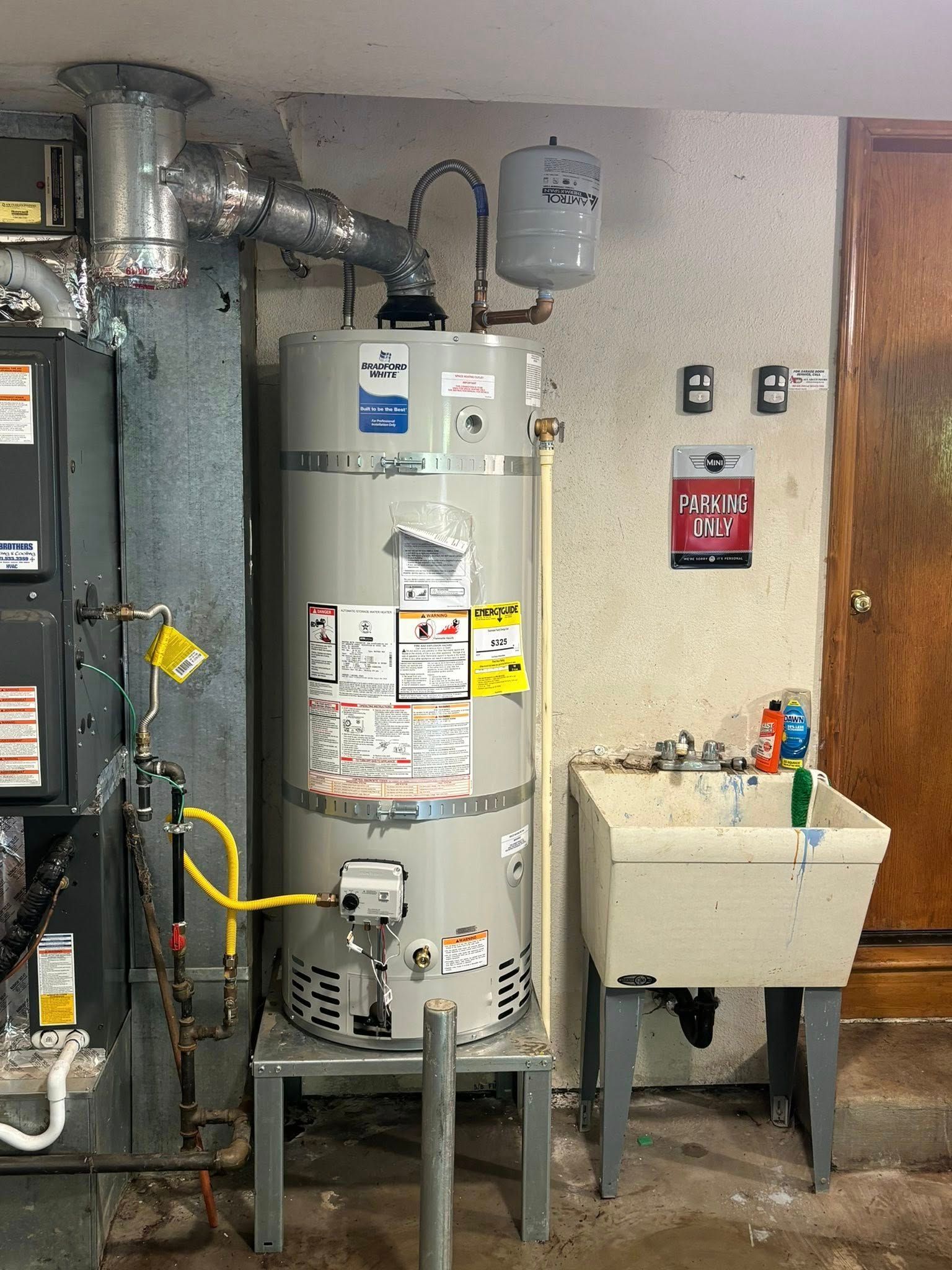 A gray water heater with an expansion tank, gas lines, and a nearby utility sink in a basement.