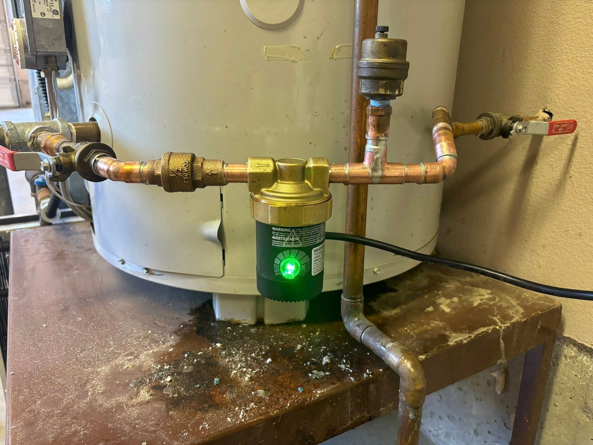 A green-lit water pump mounted on copper piping attached to the side of a water heater.