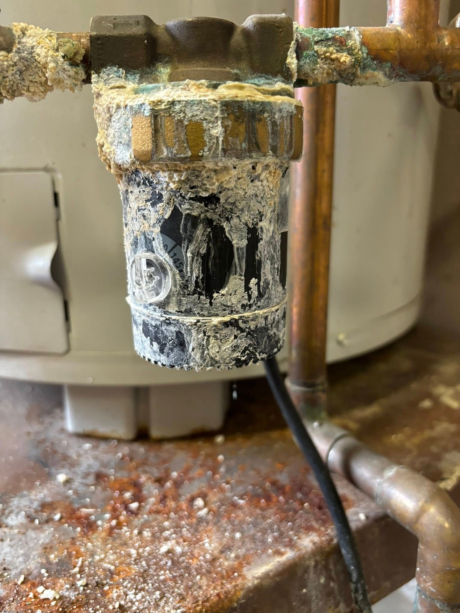 A water heater component heavily corroded with white mineral buildup and leaking onto a rusted floor.