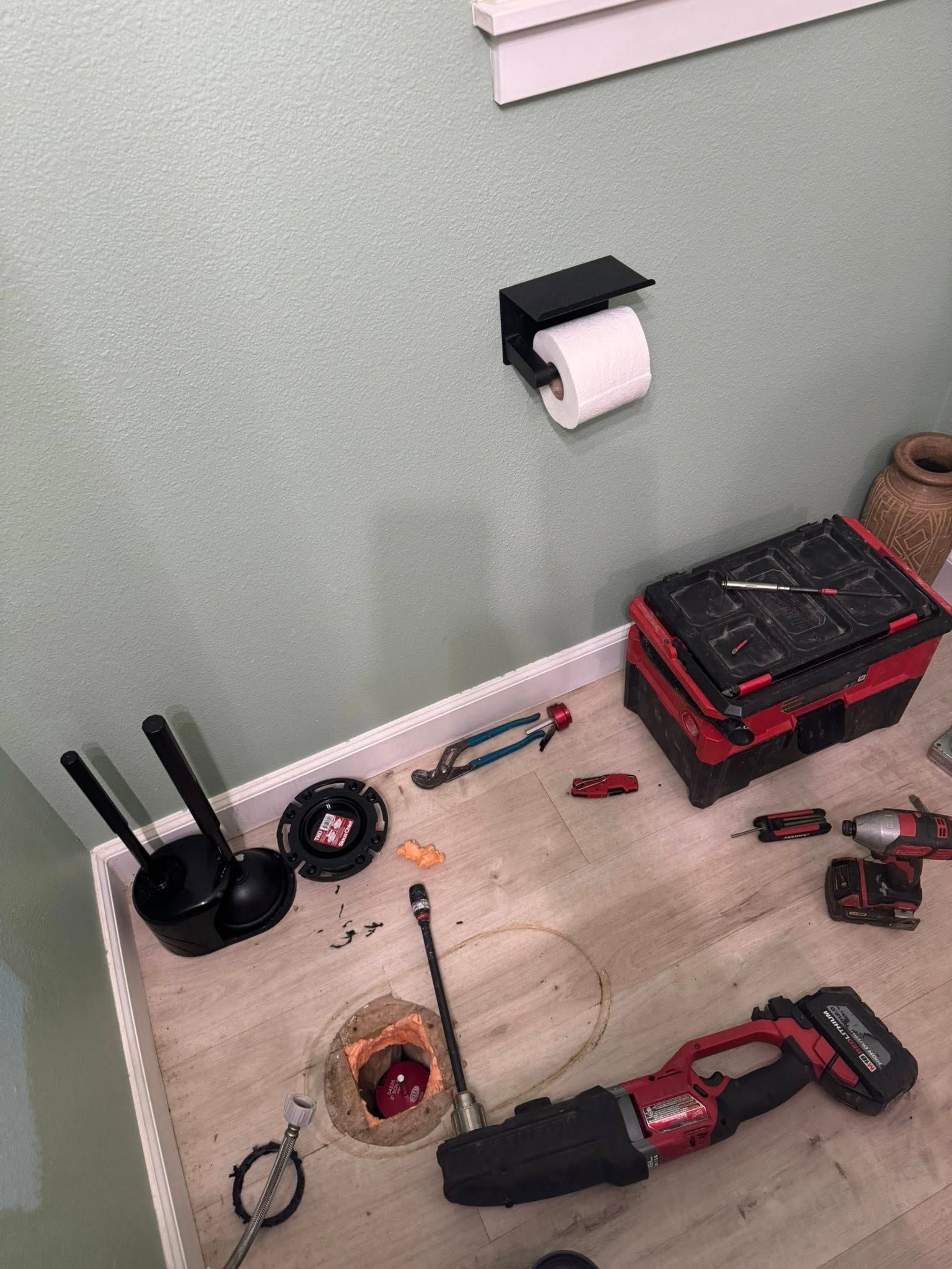 A bathroom floor under renovation with a toilet flange hole, a reciprocating saw, power tools, and a wall-mounted holder.