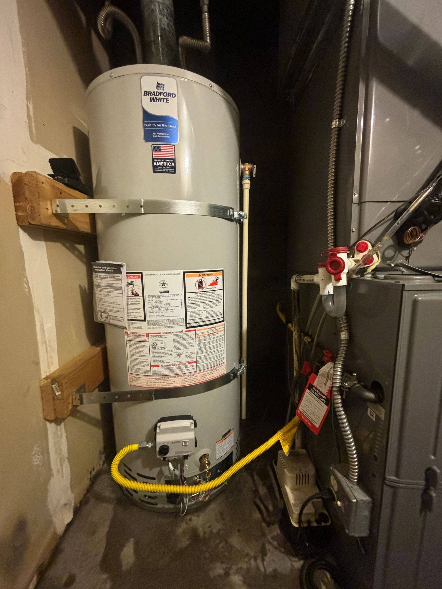 A tall, gray water heater sits next to a gray furnace in a basement, secured to the wall with metal straps.