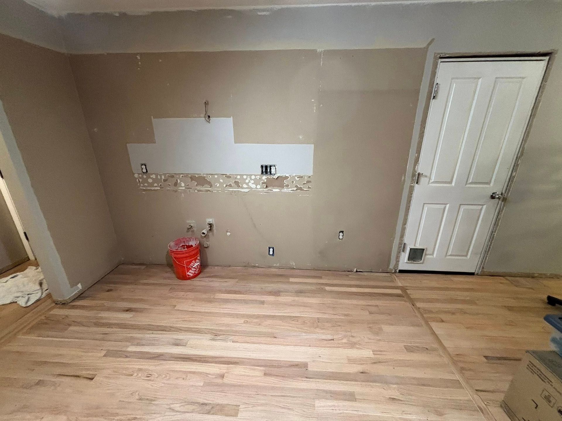 Renovated room with unfinished beige walls, exposed plumbing, a red bucket, and newly installed hardwood flooring.