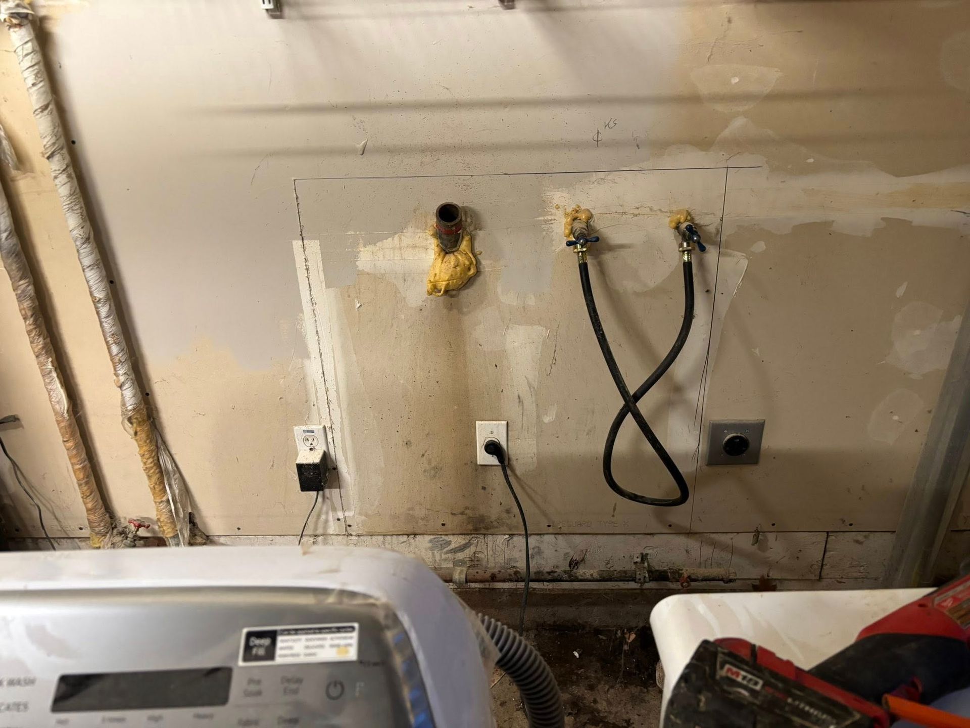 A laundry area wall with water supply valves, a drain pipe, a power outlet, and a washing machine partially in view.