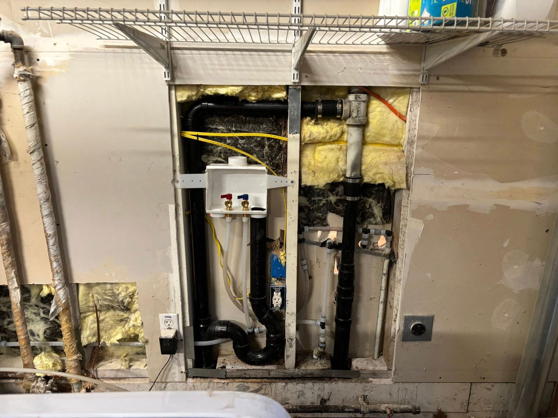 A laundry room wall with exposed framing, pipes, a washing machine outlet box, wiring, and spray foam insulation.