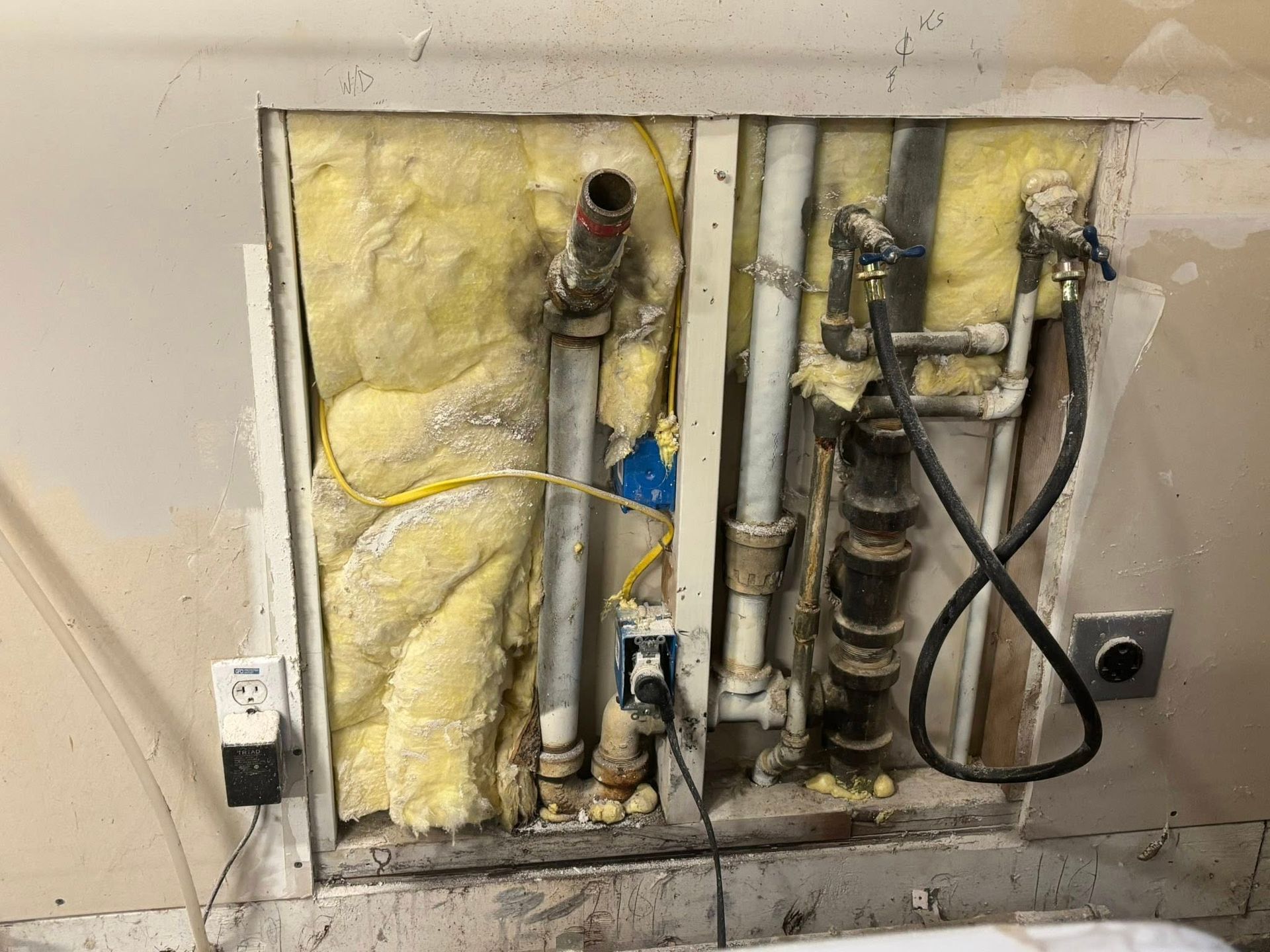 A wall section is open, revealing yellow insulation, plumbing pipes, electrical wiring, and wall outlets.