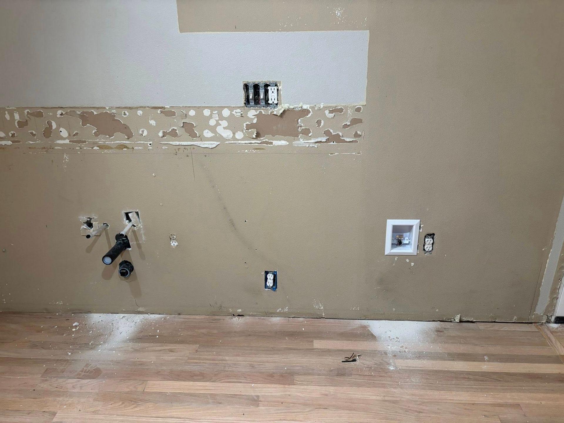 Unfinished interior wall with exposed plumbing, electrical outlets, and a recessed water supply box above wood flooring.