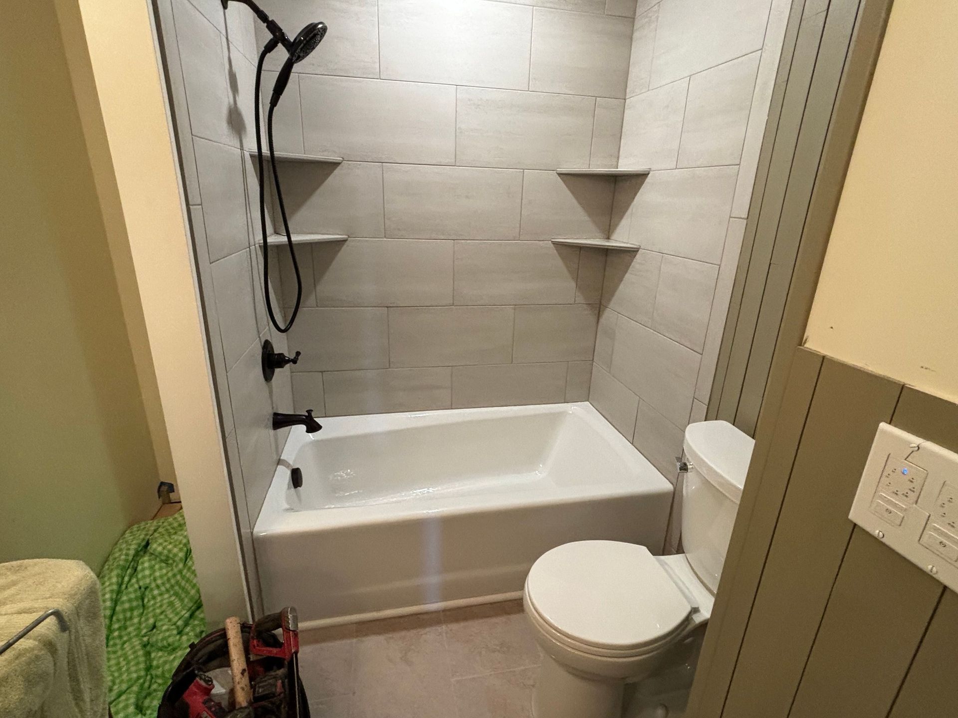 A small bathroom with a bathtub, showerhead, wall-mounted shelves, and a toilet on a light-colored tiled floor.