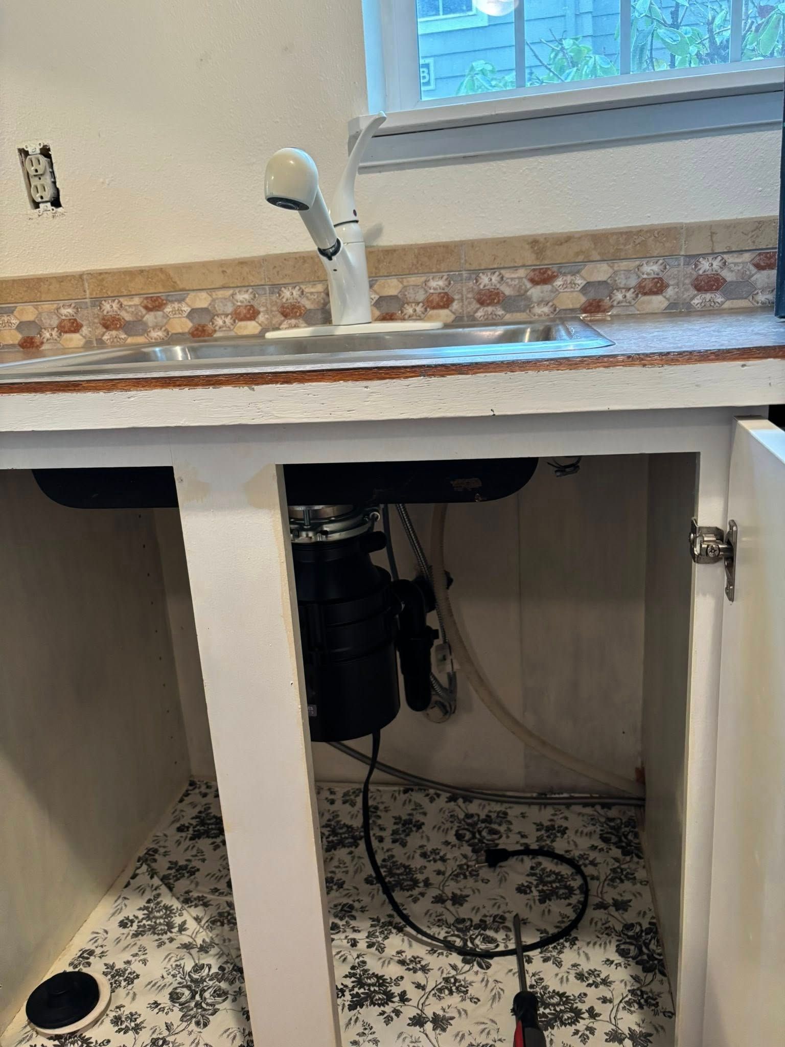 Under-sink view of a kitchen cabinet with a garbage disposal unit, plumbing, and a power cord on a patterned shelf liner.