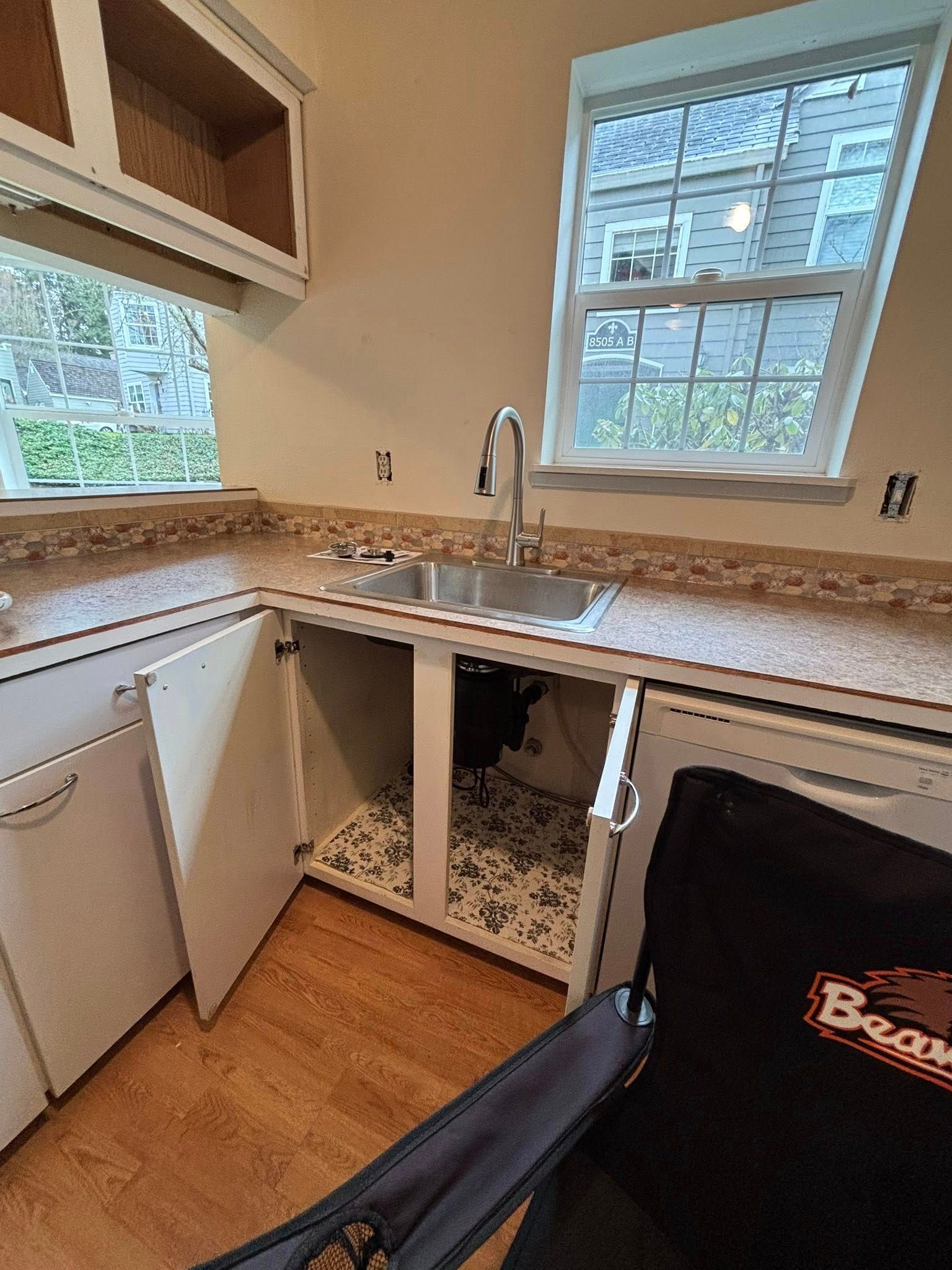 A kitchen sink area with white cabinets, a silver faucet, floral shelf lining, and a folding chair in the foreground.