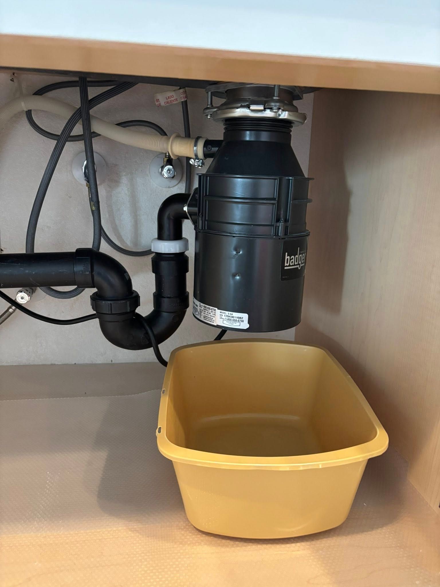 A garbage disposal unit installed under a kitchen sink, with a yellow plastic basin placed underneath it on the floor.