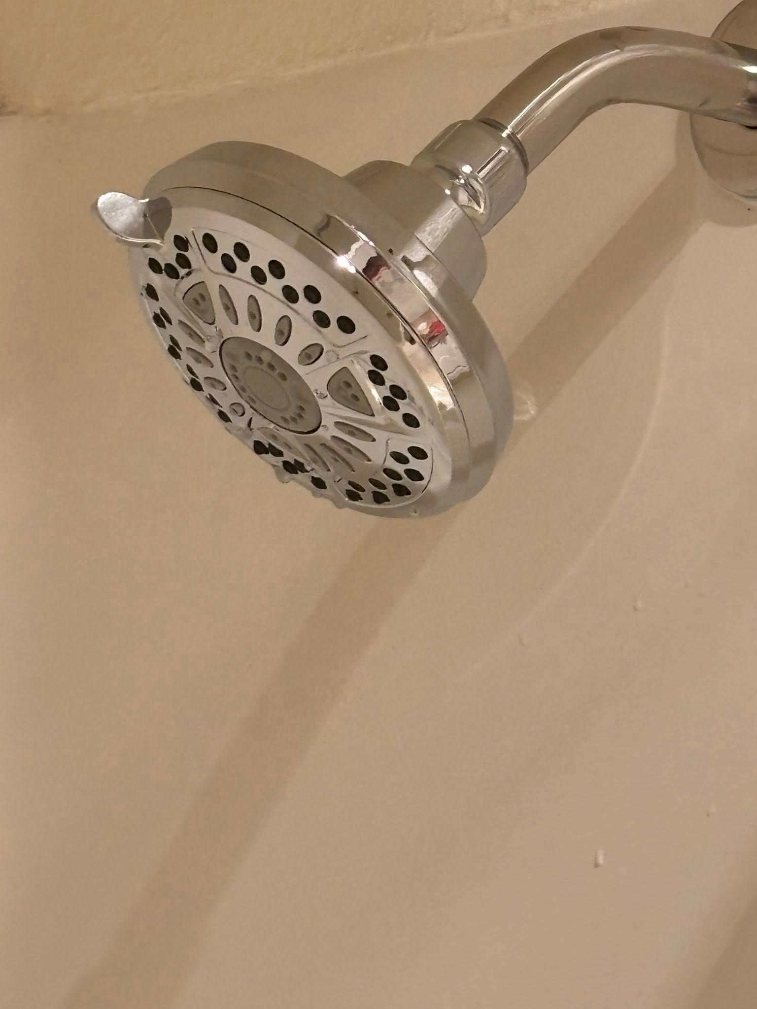 A chrome, multi-setting showerhead mounted on a shower arm, viewed from below against a beige tiled wall.