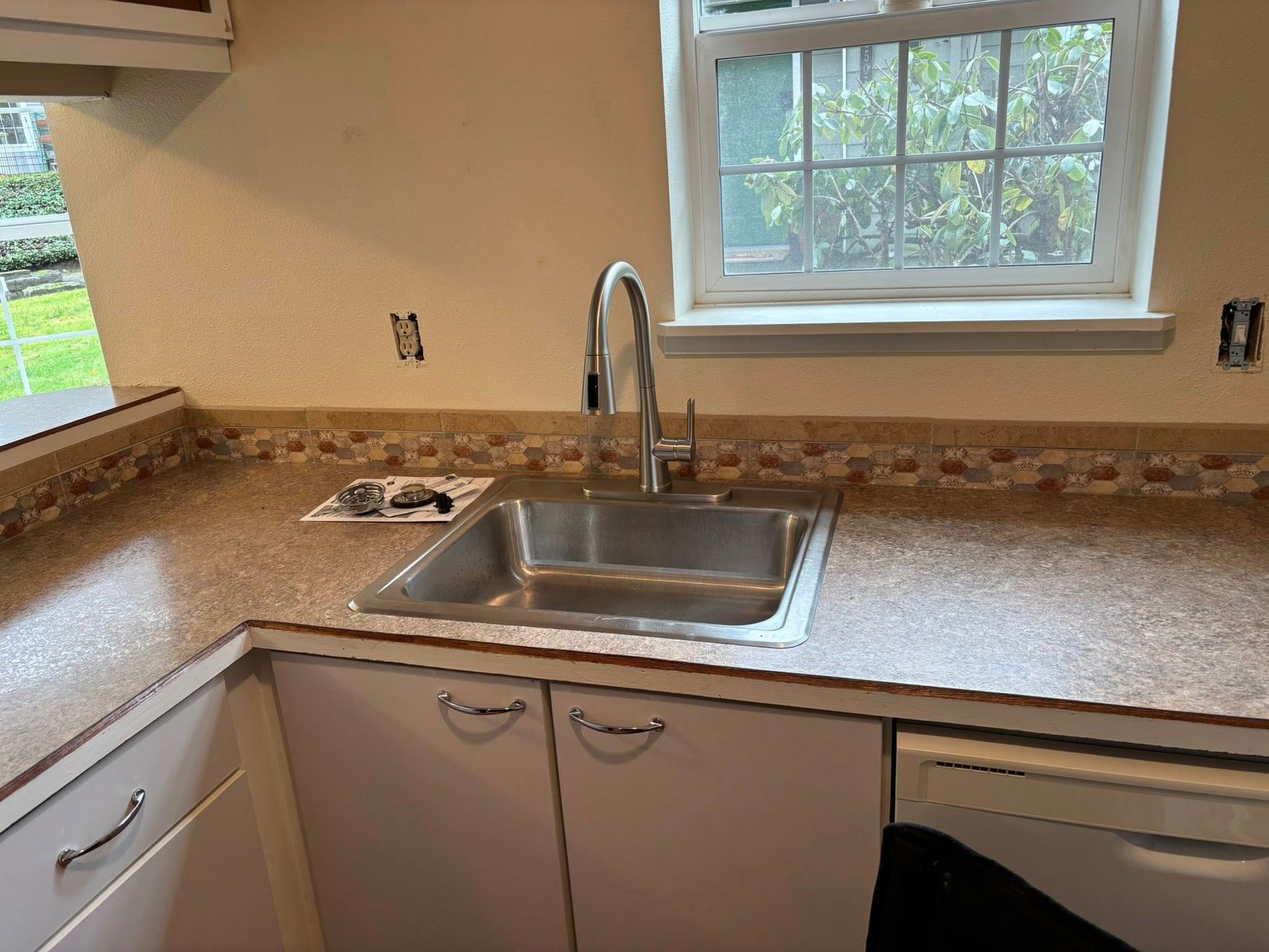 A kitchen sink with a stainless steel faucet and basin set in a tan, speckled countertop below a window.