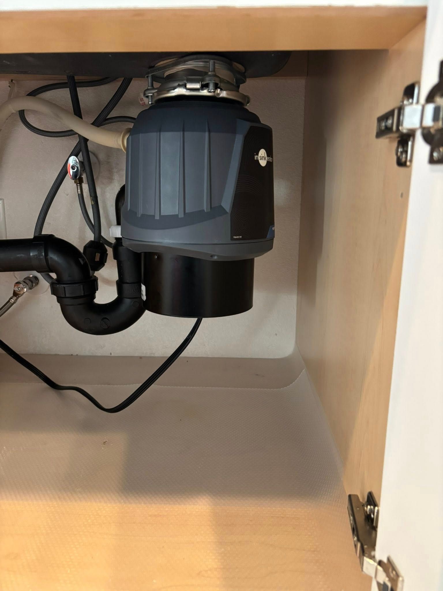 A gray and black garbage disposal installed under a kitchen sink, with black plumbing pipes visible to the left.