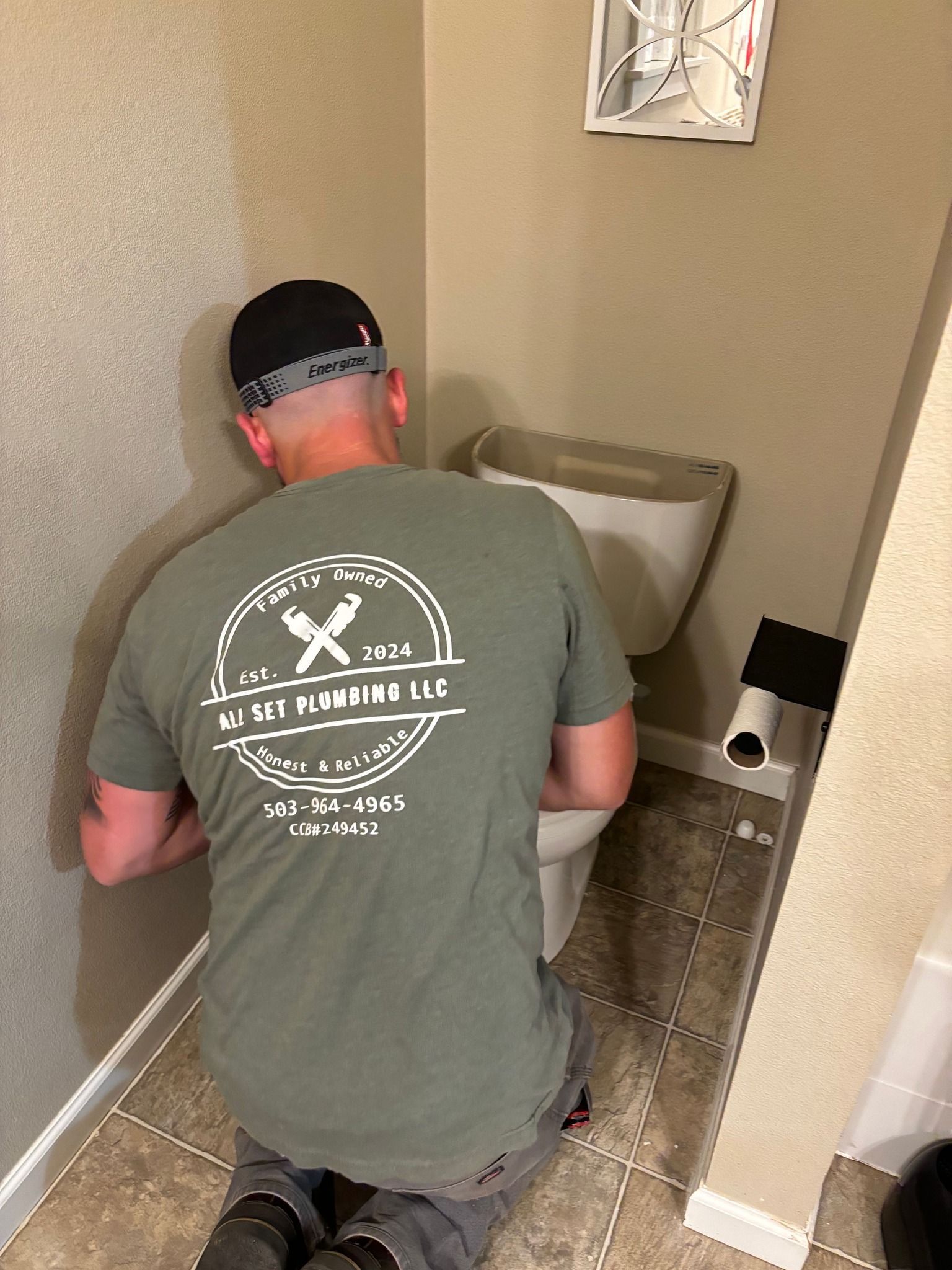 A person wearing a green work t-shirt kneels on a bathroom floor while repairing a toilet tank.